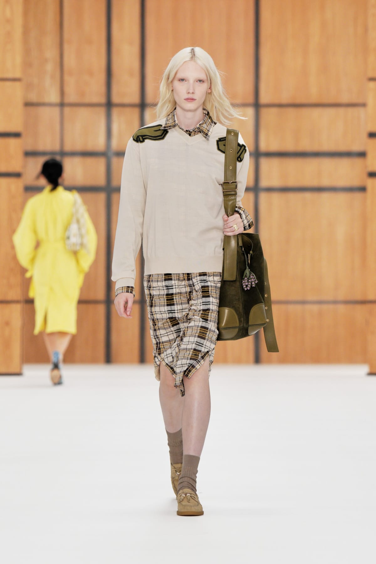 A model on the Burberry Spring/Summer 2027 runway in a beige plaid skirt and cream sweater with olive shoulder patches, carrying a large olive suede and leather bag