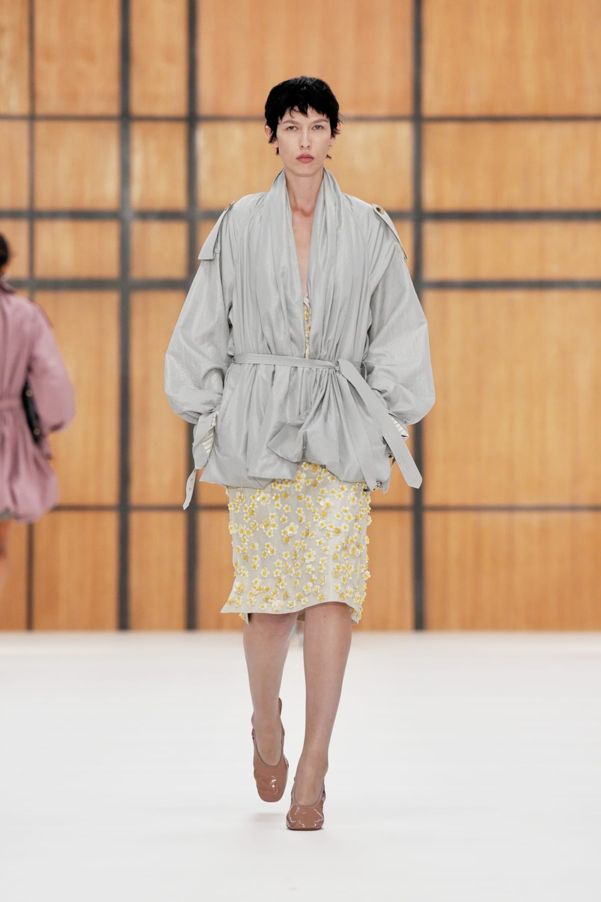 A model on the Burberry Spring/Summer 2027 runway in a grey wrap jacket over a cream and yellow floral-embroidered pencil skirt
