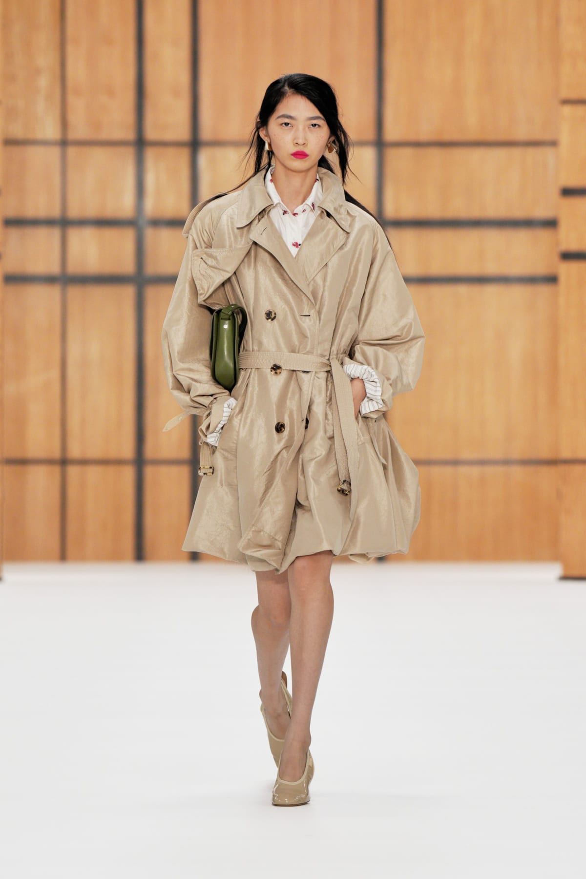 A model on the Burberry Spring/Summer 2027 runway in a beige trench coat with a bubble hem and belted waist, carrying an olive-green bag