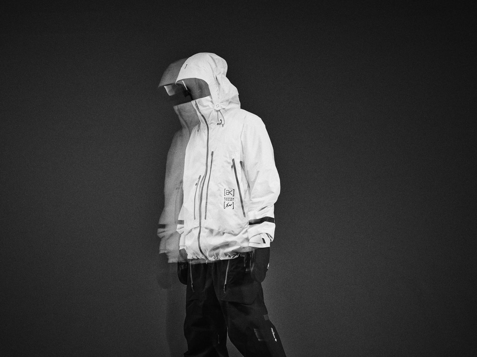 A model in a white hooded Burton x fragment shell jacket and black pants against a dark background, shot with motion blur, the [ak], Burton and fragment marks stacked on the front between the zips