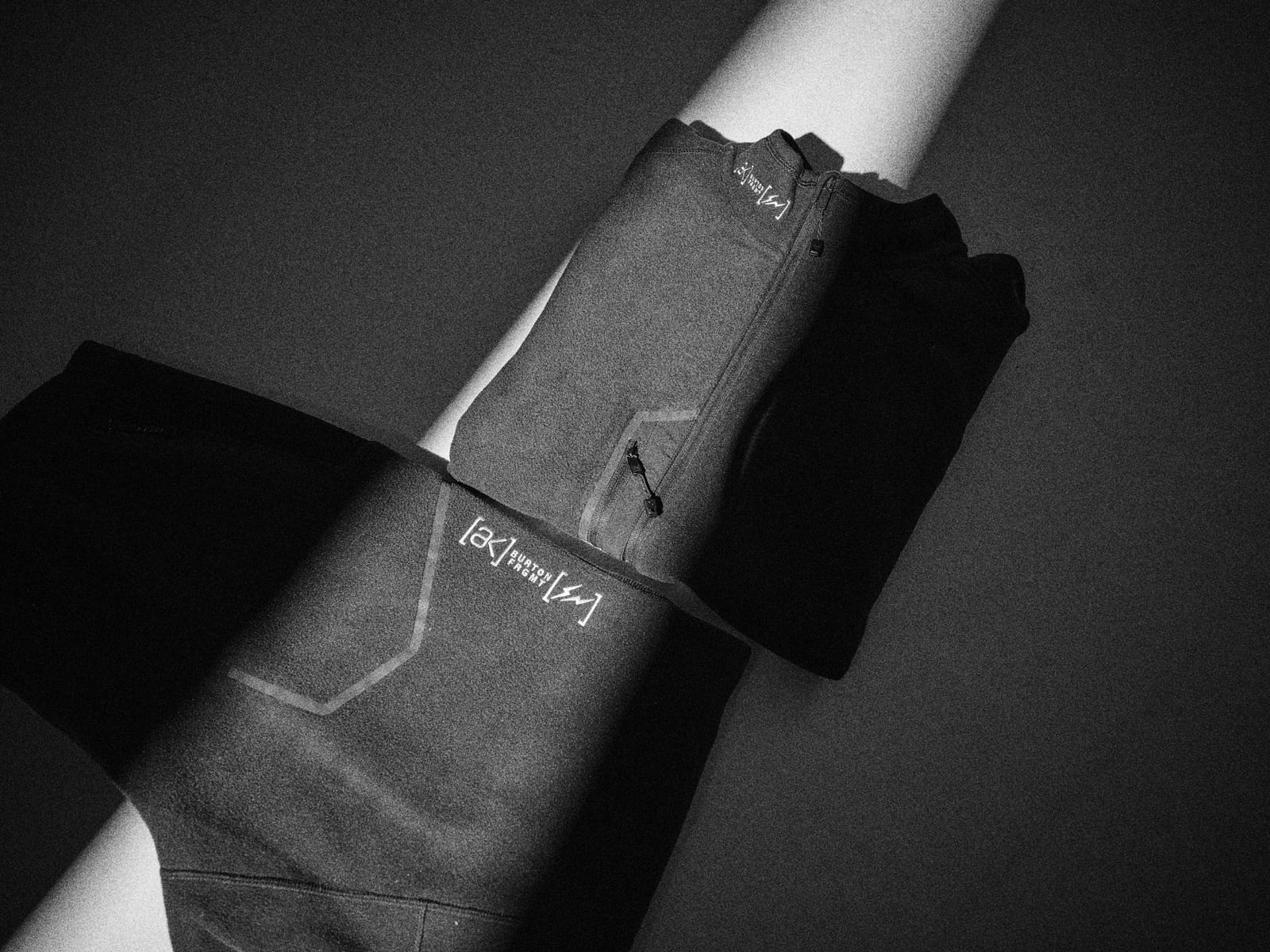 A dark Burton x fragment zip fleece and fleece pants laid across a strip of light, each printed with the [ak] and fragment marks