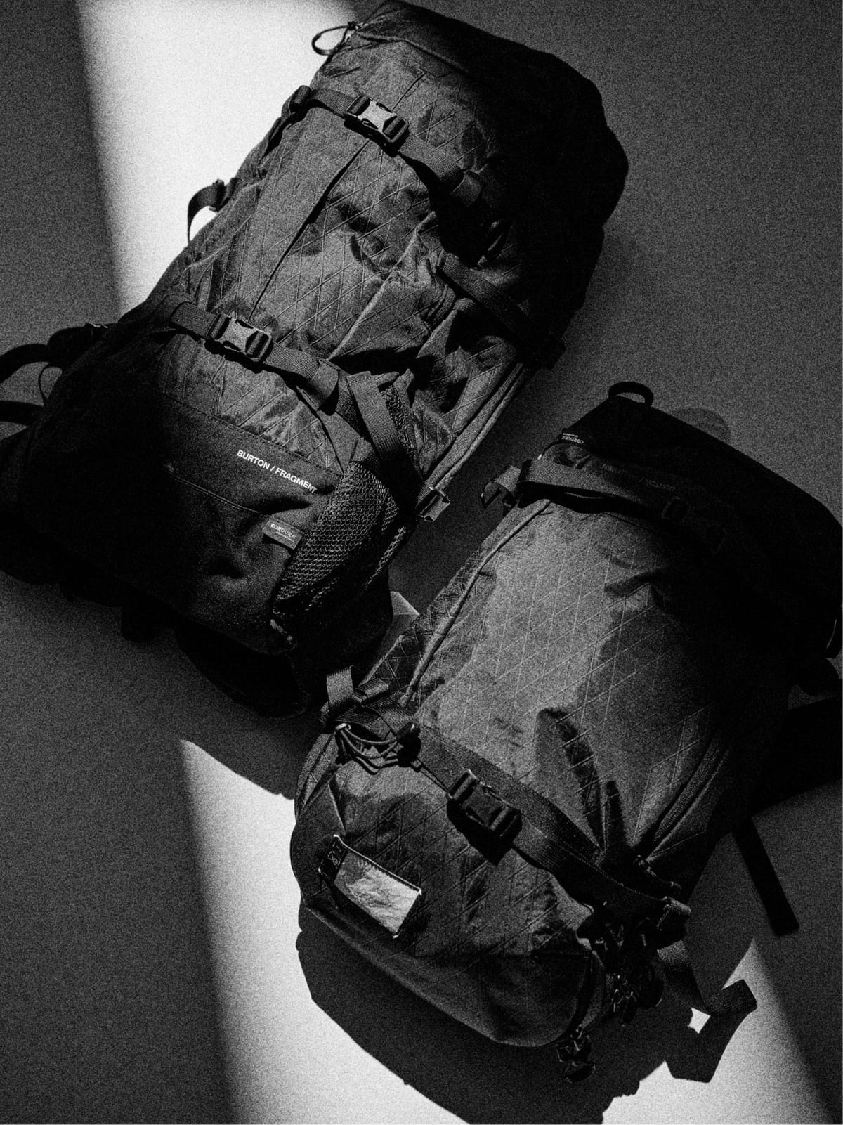 Two black Burton x fragment backpacks in a gridded ripstop lying in a strip of light, one with a BURTON / FRAGMENT label