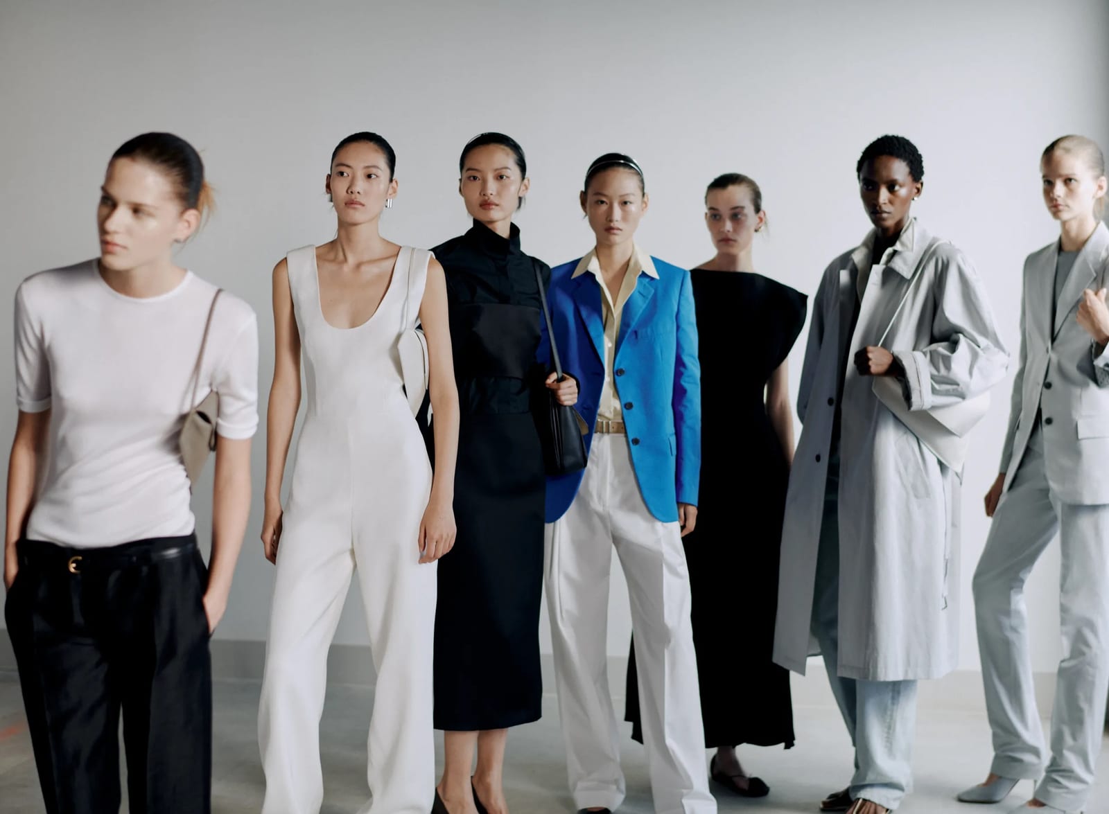 Seven models in Calvin Klein Collection's Spring 2027 looks, lined up against a pale wall in a white T-shirt and black trousers, a white jumpsuit, a black dress, a bright blue blazer with white trousers, a black dress, a long grey coat and a pale grey suit