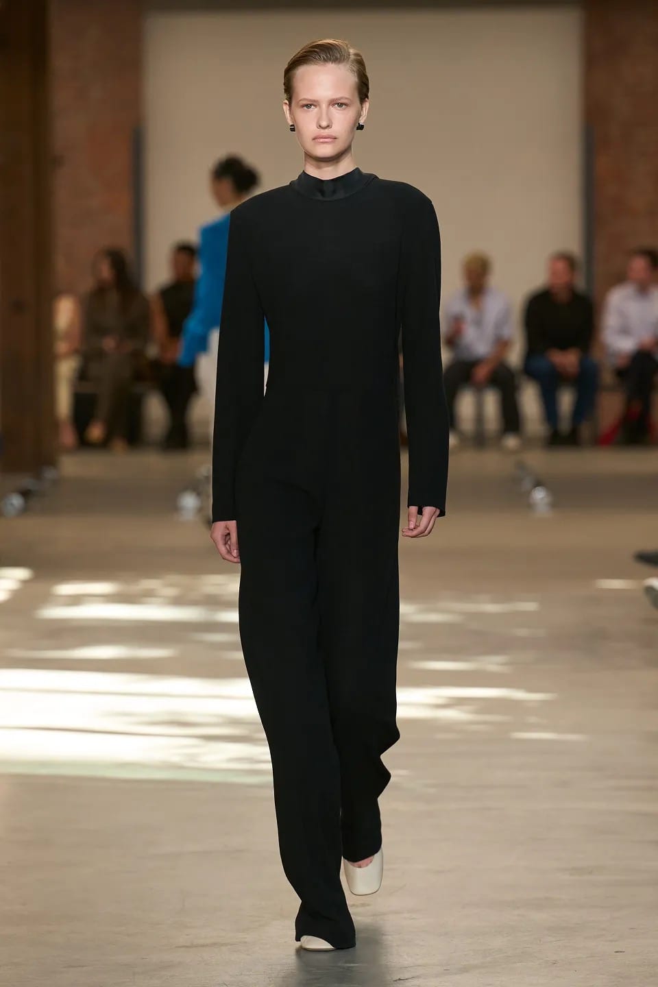 A model with slicked-back hair walking the Spring 2027 runway in an all-black long-sleeved high-neck look with wide trousers and white shoes, past a seated front row in a brick warehouse