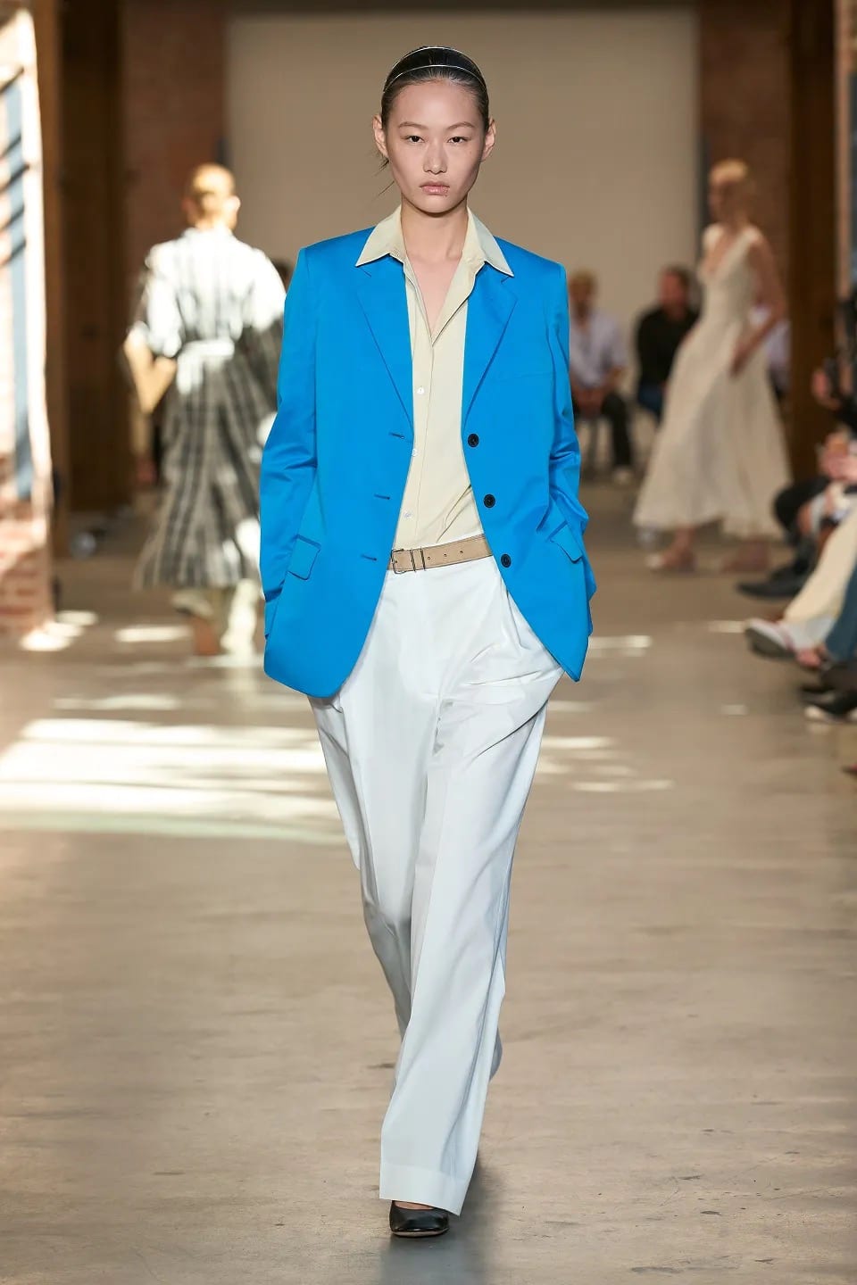 A model on the Spring 2027 runway at Terminal Warehouse in a bright blue blazer over a pale yellow shirt, wide white trousers and a tan belt, with other models and seated guests behind her