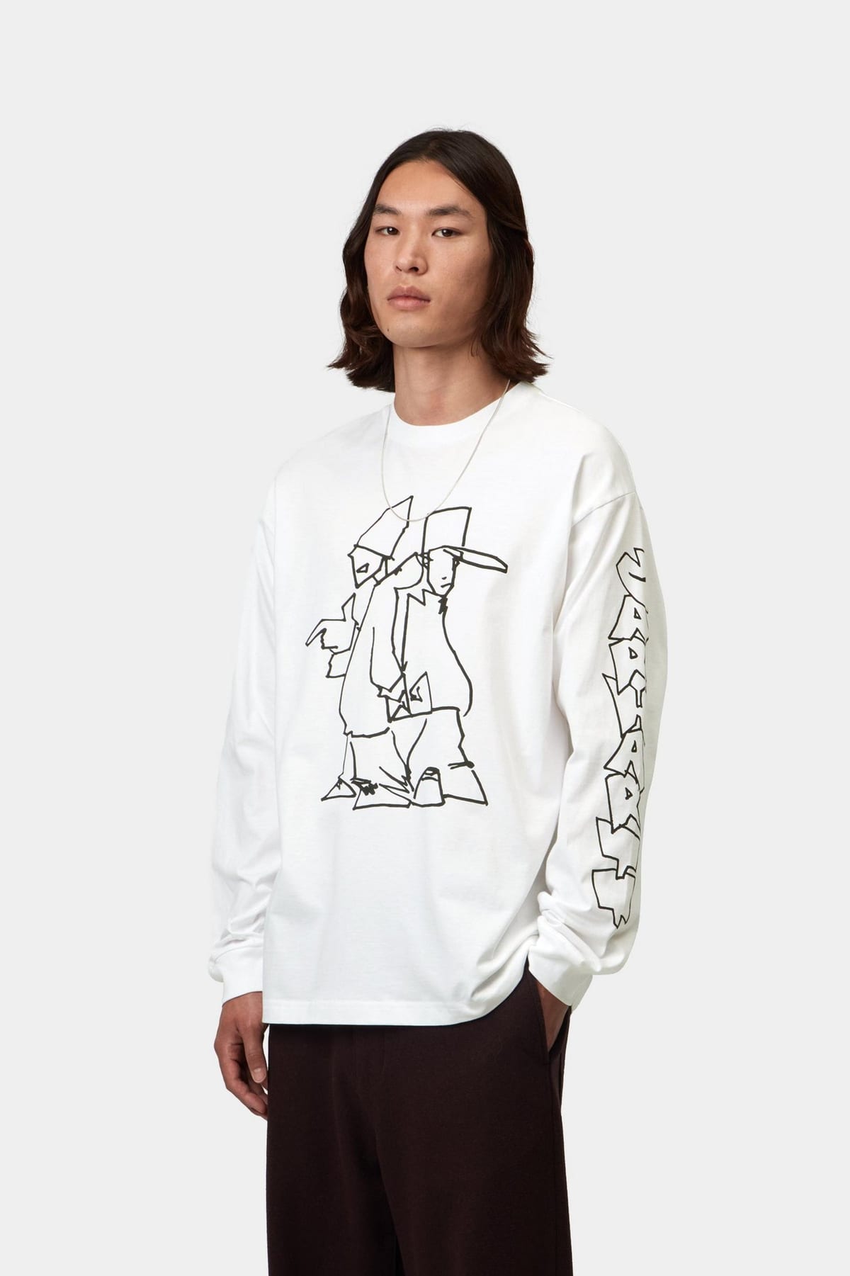 A man with shoulder-length dark hair in a white long-sleeve Carhartt WIP T-shirt printed with Blinkerfluid's black line drawing of two figures in tall hats, one pointing, and blocky graffiti lettering running down the left sleeve, with dark brown trousers and a thin silver chain