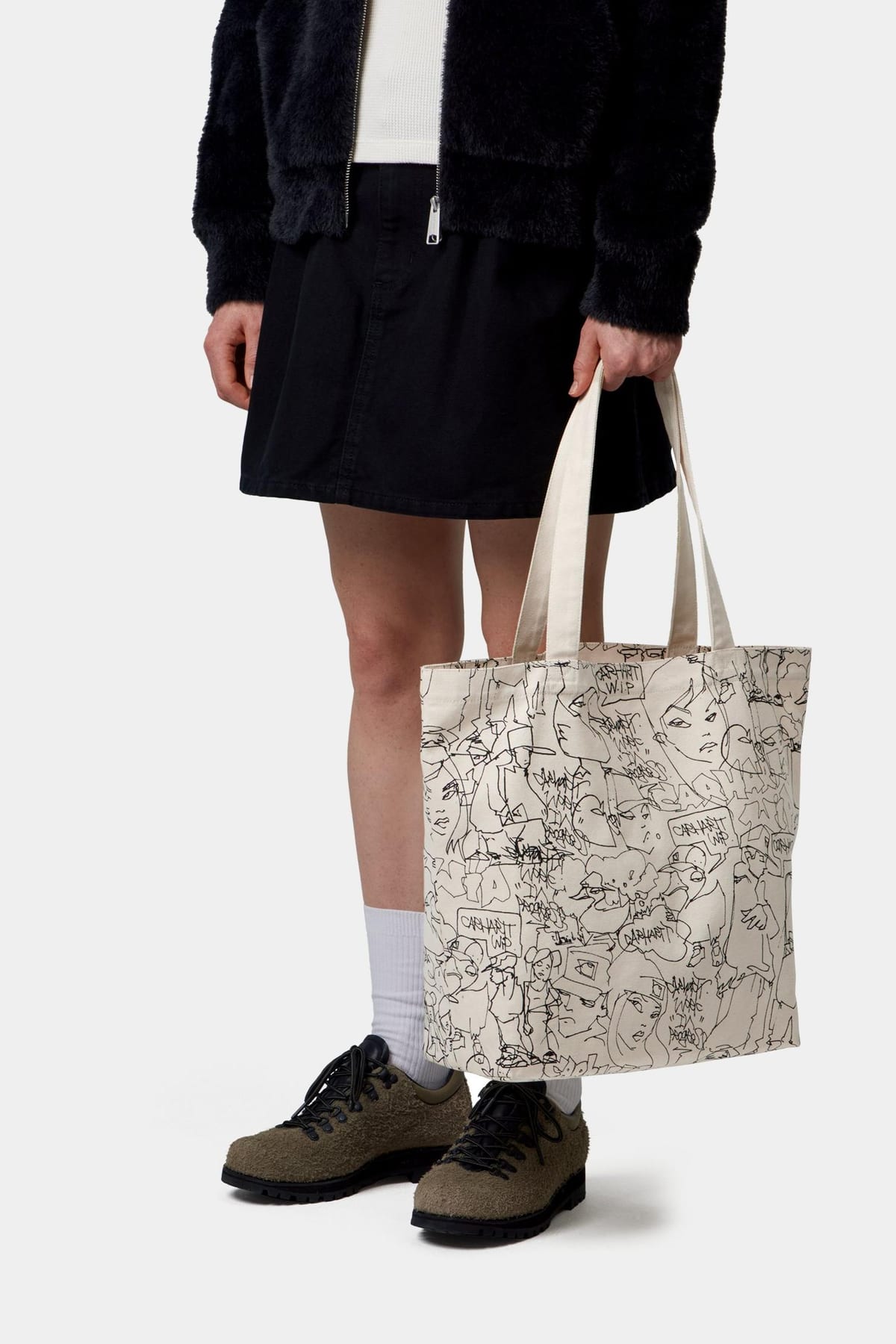 A person in a black fuzzy zip jacket, a black denim skirt, white socks and olive suede hiking shoes holding a natural canvas tote covered all over in black line drawings of faces and figures, a duck among them, with 'Carhartt W.I.P.' written in speech bubbles
