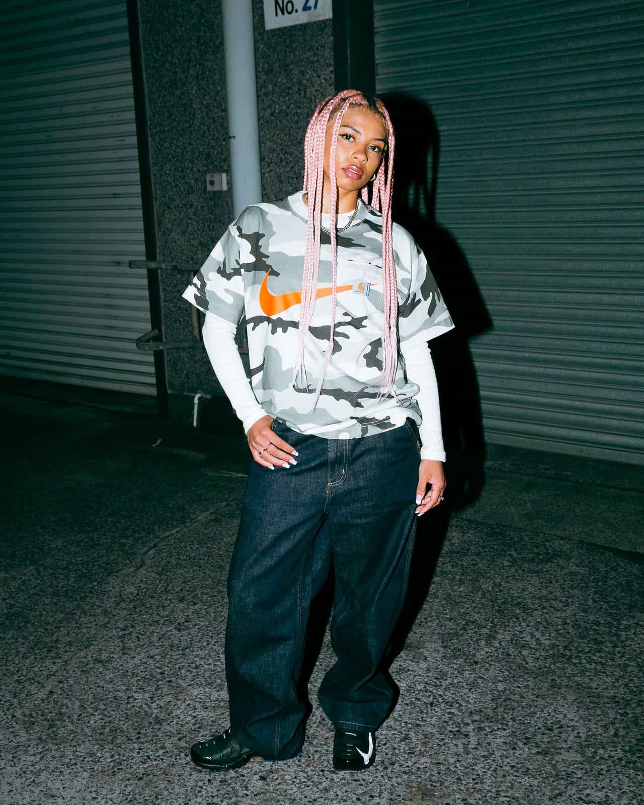 A person with long pink braids in a grey and white camouflage T-shirt with an orange swoosh and a Carhartt WIP label on the chest, over a white long-sleeve and dark baggy jeans