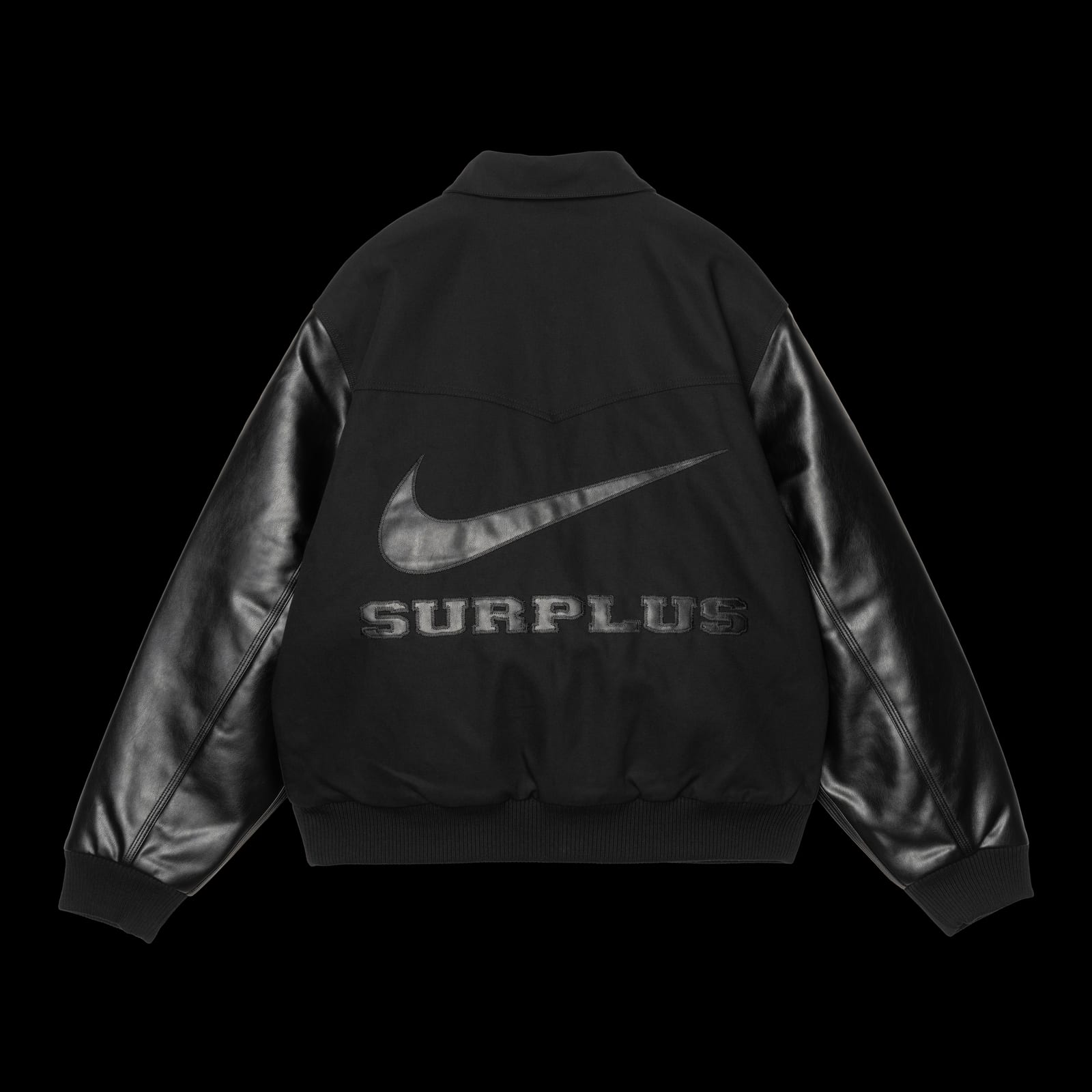 The back of a black jacket with faux-leather sleeves, a black swoosh appliqué and SURPLUS lettered in a collegiate typeface beneath it