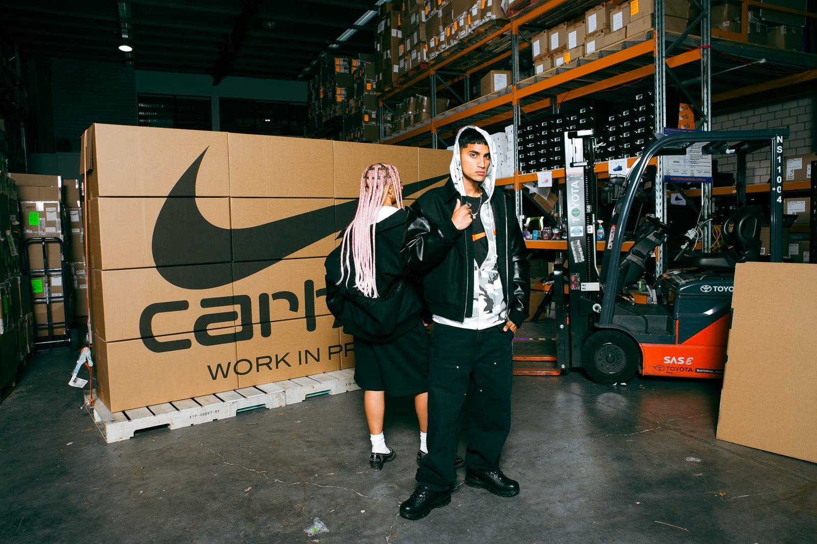 Two people posed in a warehouse in front of a stack of oversized boxes printed with the Nike swoosh and Carhartt WIP name: one in a black canvas jacket with black sleeves over a white camouflage hoodie, the other in a black hooded jacket seen from behind