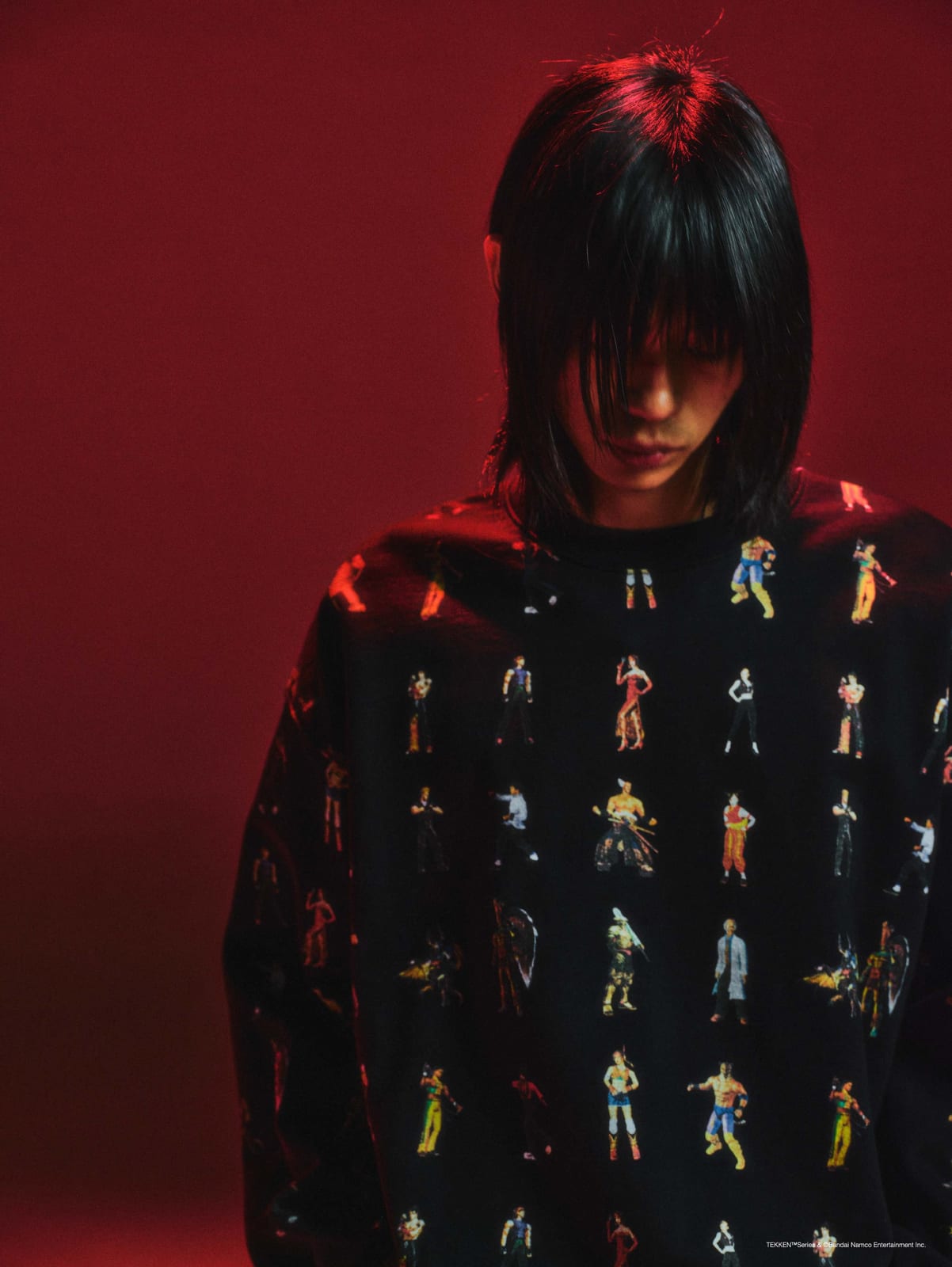 A model in the black Carnival x Tekken long-sleeve top printed with rows of Tekken characters, against a red backdrop