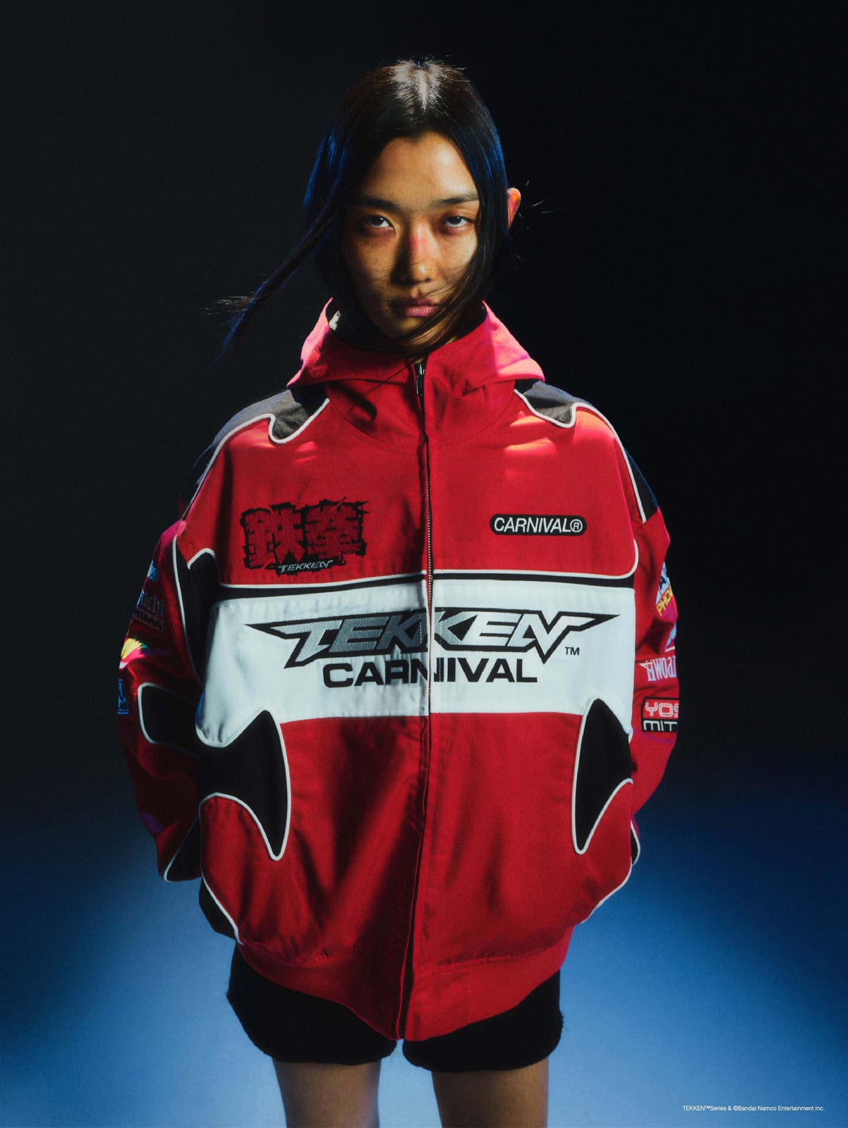 A model in the red and white Carnival x Tekken racing jacket, with TEKKEN and CARNIVAL across a white chest panel and 鉄拳 patched on the chest