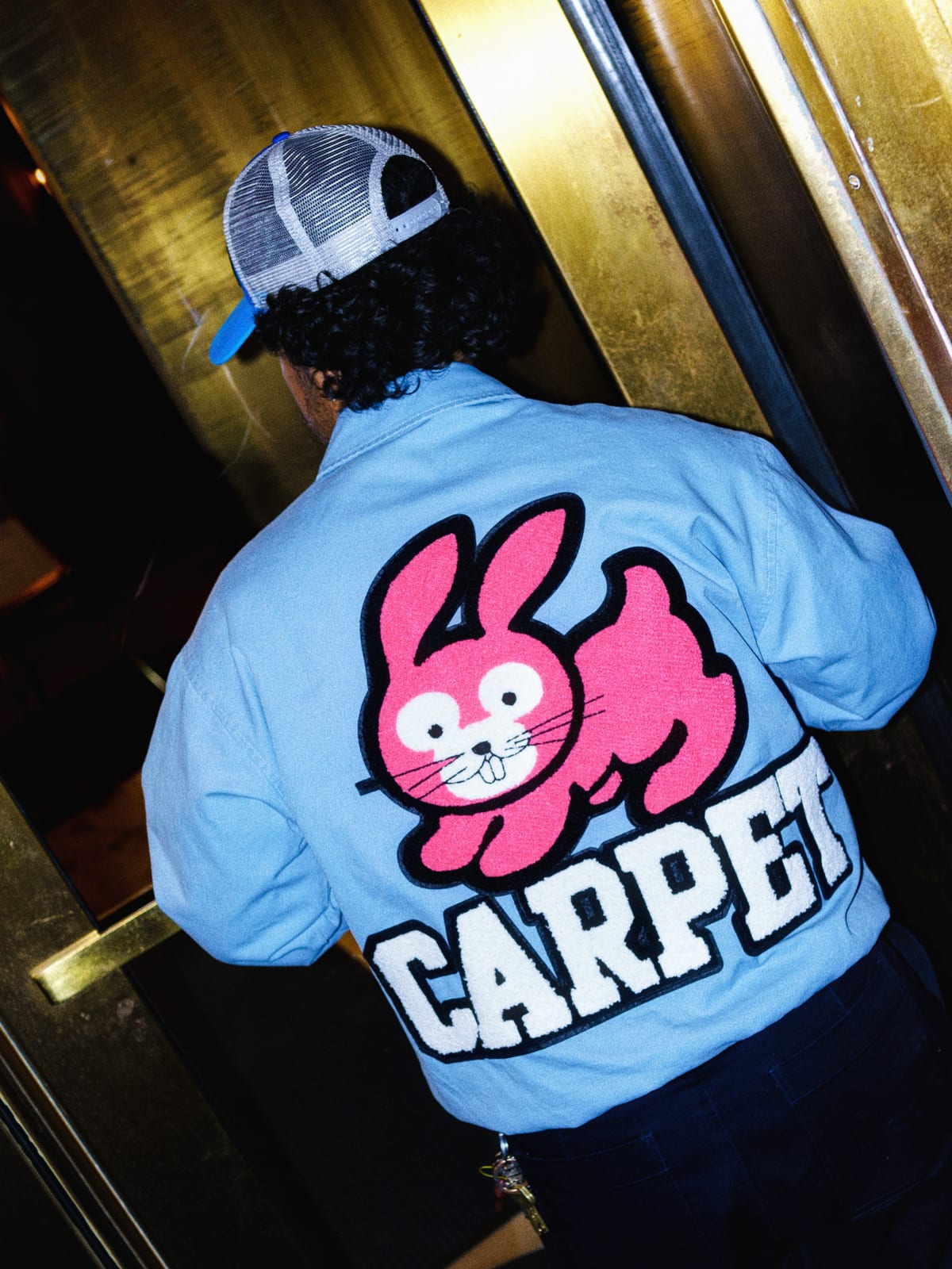 The back of a person in a light blue canvas work jacket with a large pink chenille cartoon rabbit above white CARPET lettering, wearing a trucker cap backwards
