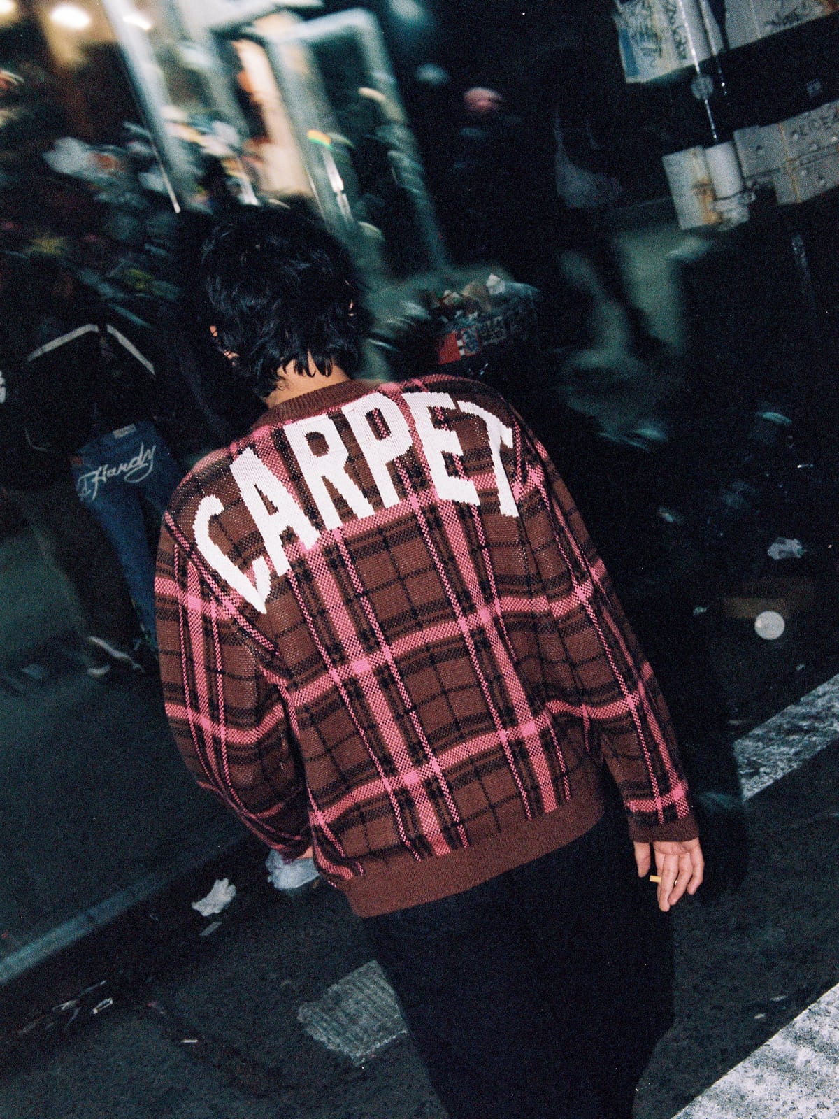 A man seen from behind on a busy street at night in a brown and pink plaid knit sweater with CARPET in large white letters across the shoulders