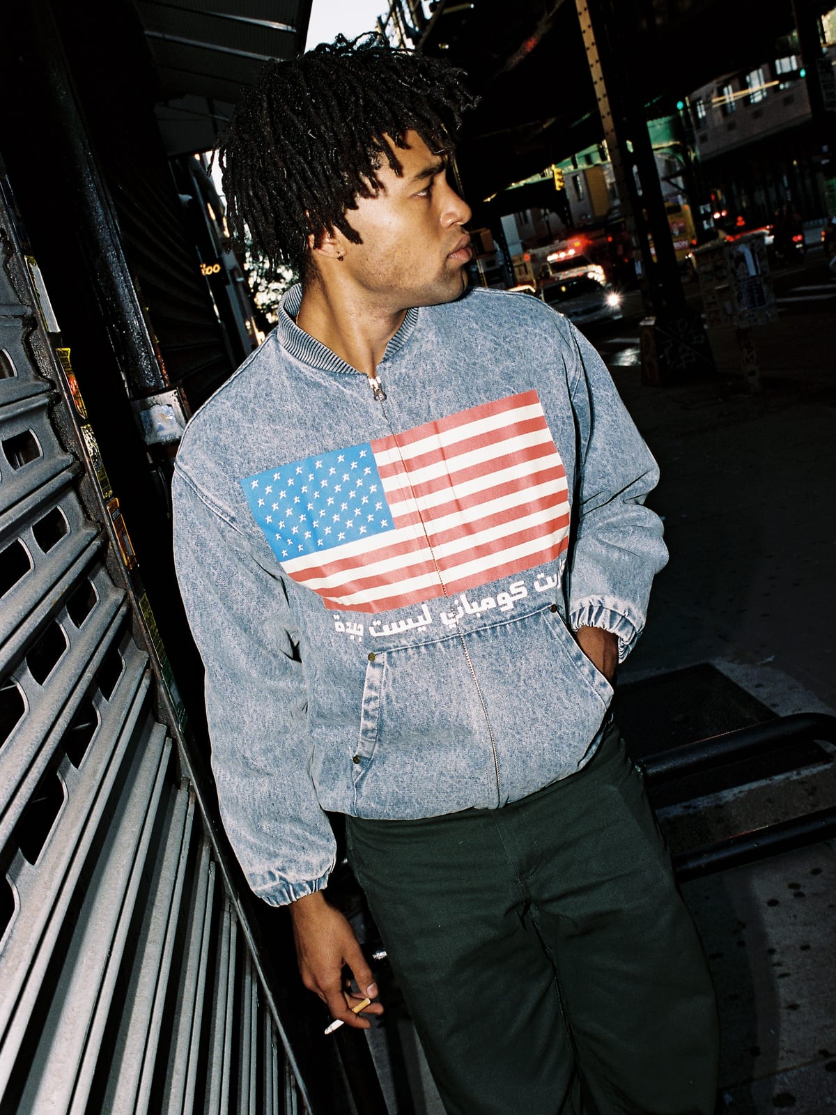 A man with twisted locs on a city street under an elevated train line at dusk in a light acid-wash denim bomber jacket printed with a large American flag across the chest and a line of Arabic lettering under it, with dark green trousers