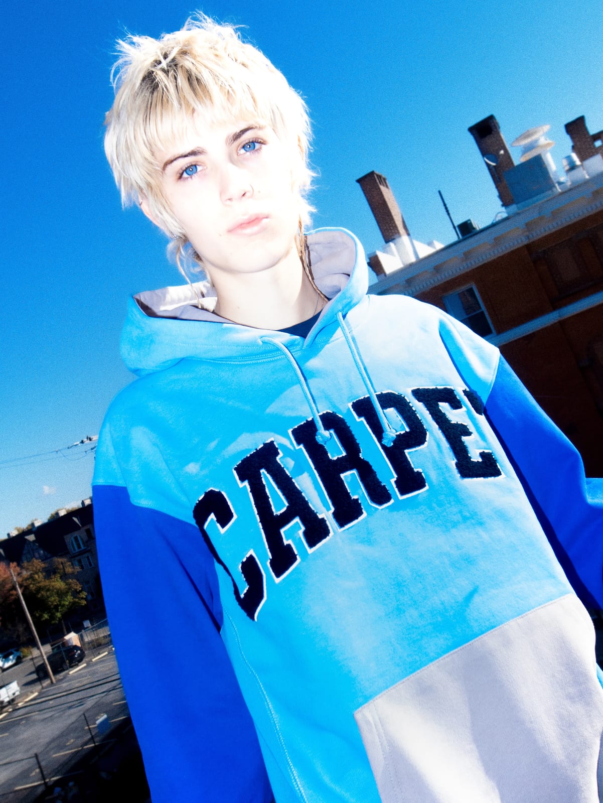 A person with bleached hair against a bright blue sky in a hoodie colour-blocked in light blue, royal blue and grey, with CARPET in black chenille arch lettering across the chest