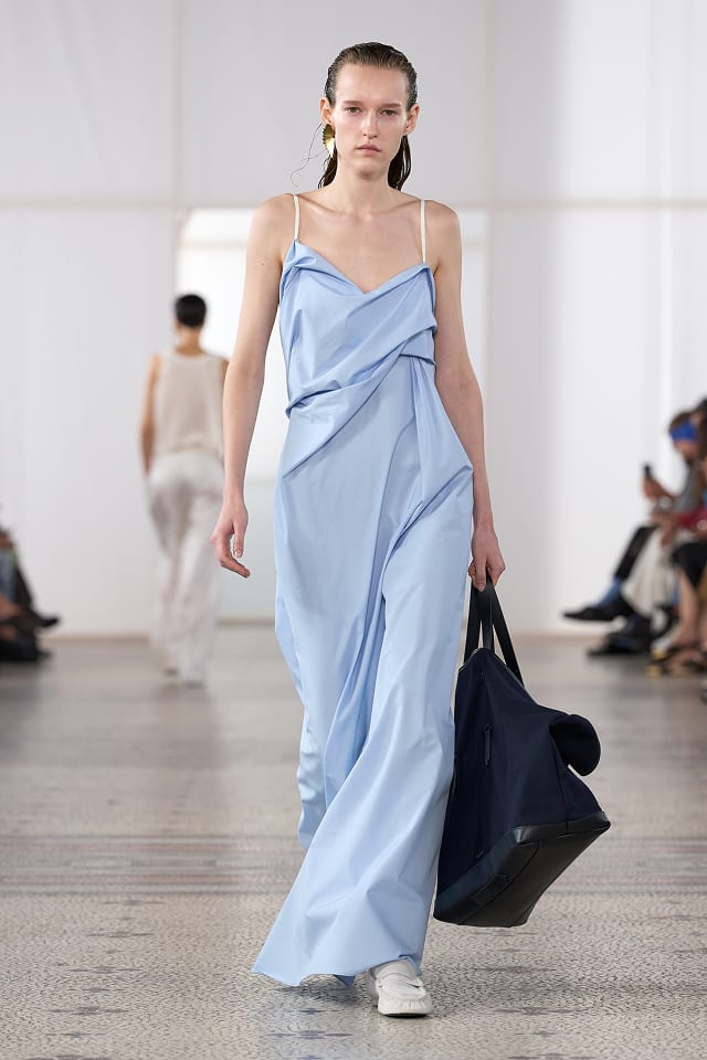 A model in a pale blue draped maxi dress with thin straps, carrying a dark tote bag
