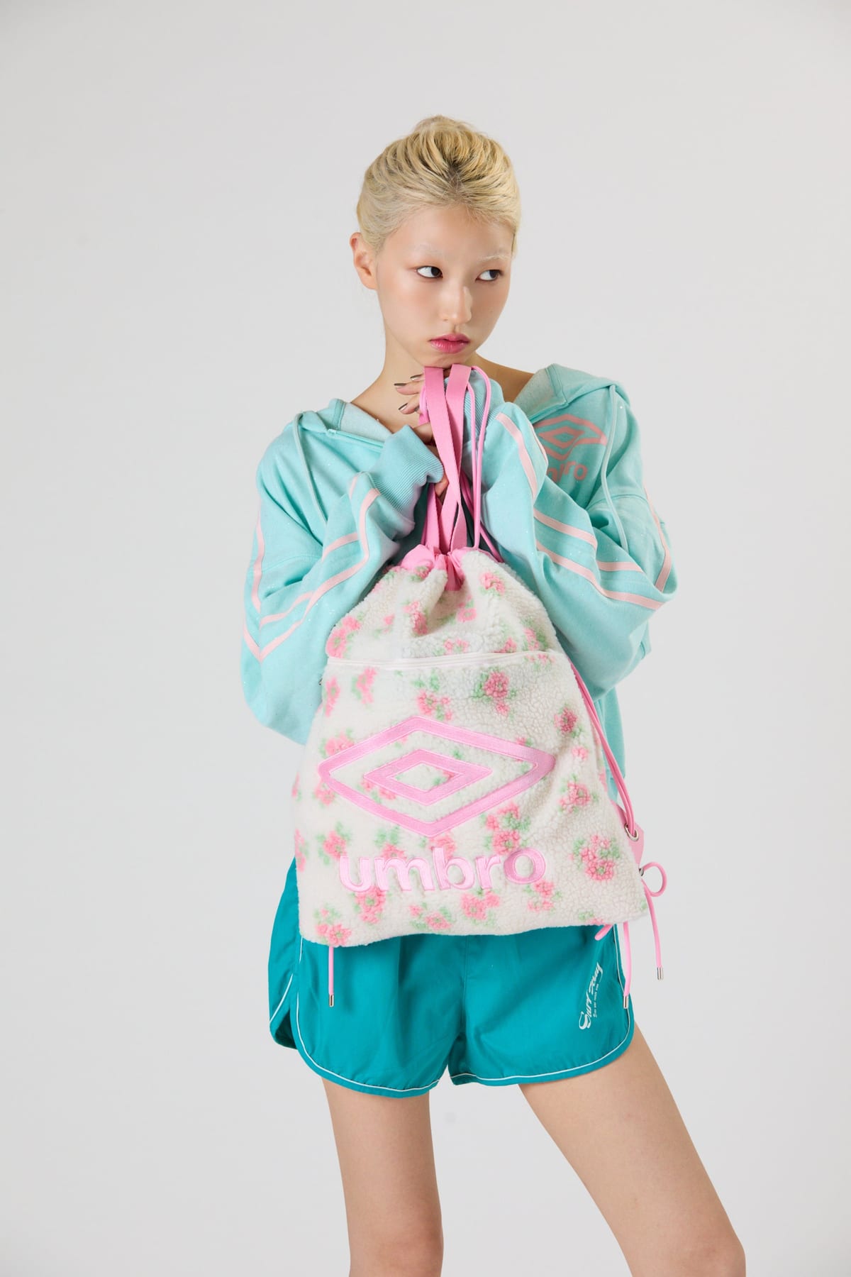 A model in a mint-green zip hoodie holding a cream drawstring knapsack with a pink floral pattern and a pink Umbro logo