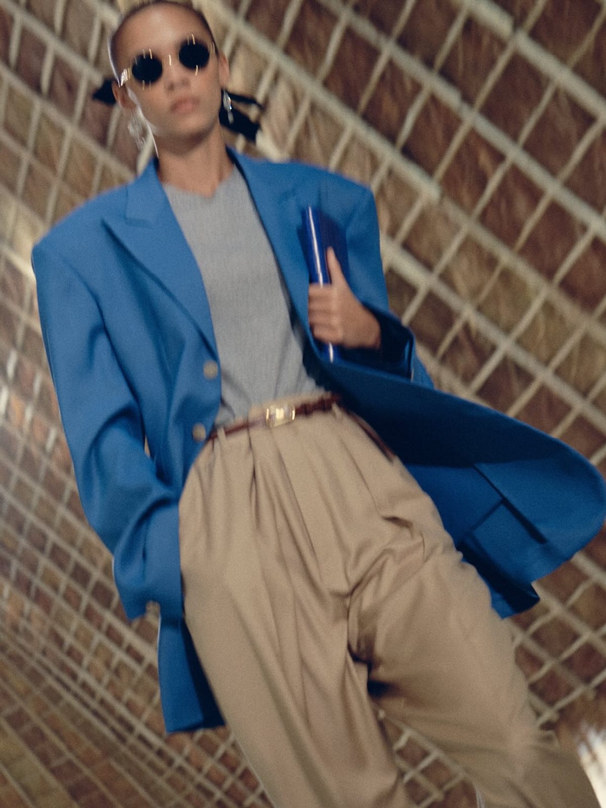 A Celine Spring 2027 look: a bright blue oversized blazer over a grey T-shirt and pleated tan trousers with a thin belt, worn with small round sunglasses