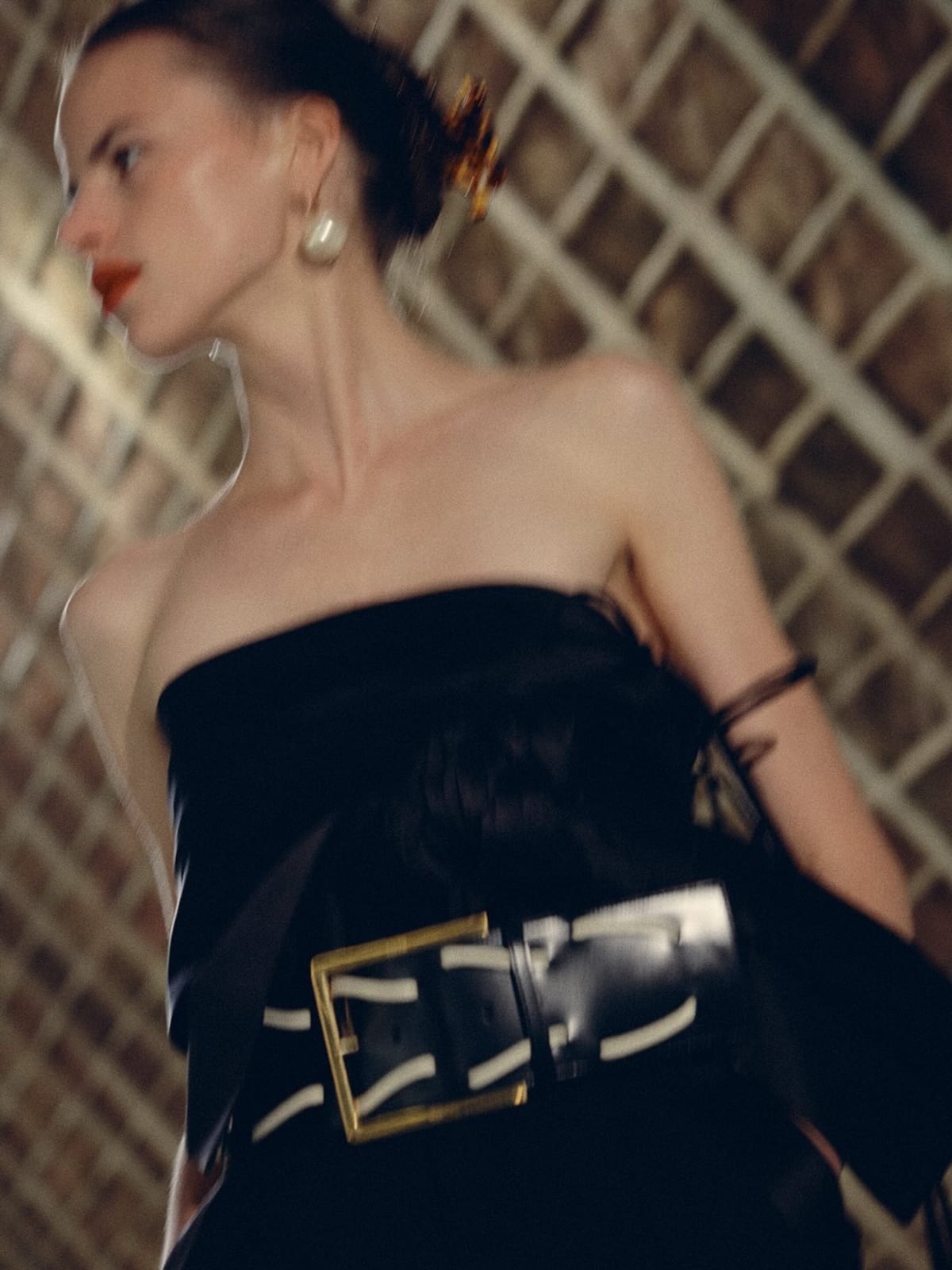 A Celine Spring 2027 look: a black strapless dress cinched with an oversized black leather belt and a large gold buckle, worn with pearl earrings