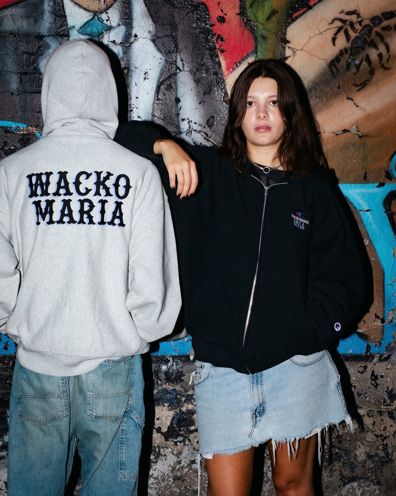 Two models against a graffiti wall: one with their back turned in a grey hoodie with WACKO MARIA in black letters across the back, the other in a black zip hoodie with a small embroidered chest logo and a frayed denim skirt