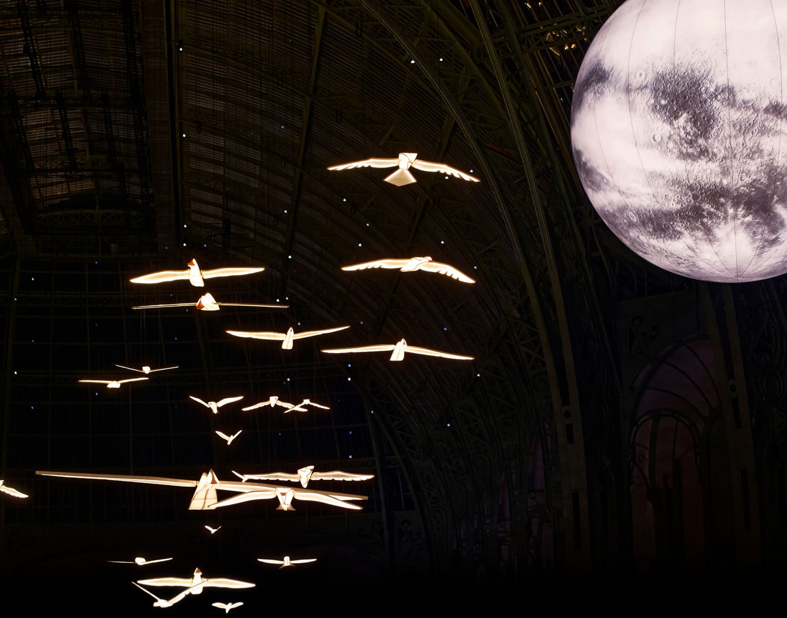 The Chanel Spring 2027 set inside the Grand Palais: a lit white moon at the top right of a dark glass-and-steel dome, with a flock of glowing white birds in flight beneath it