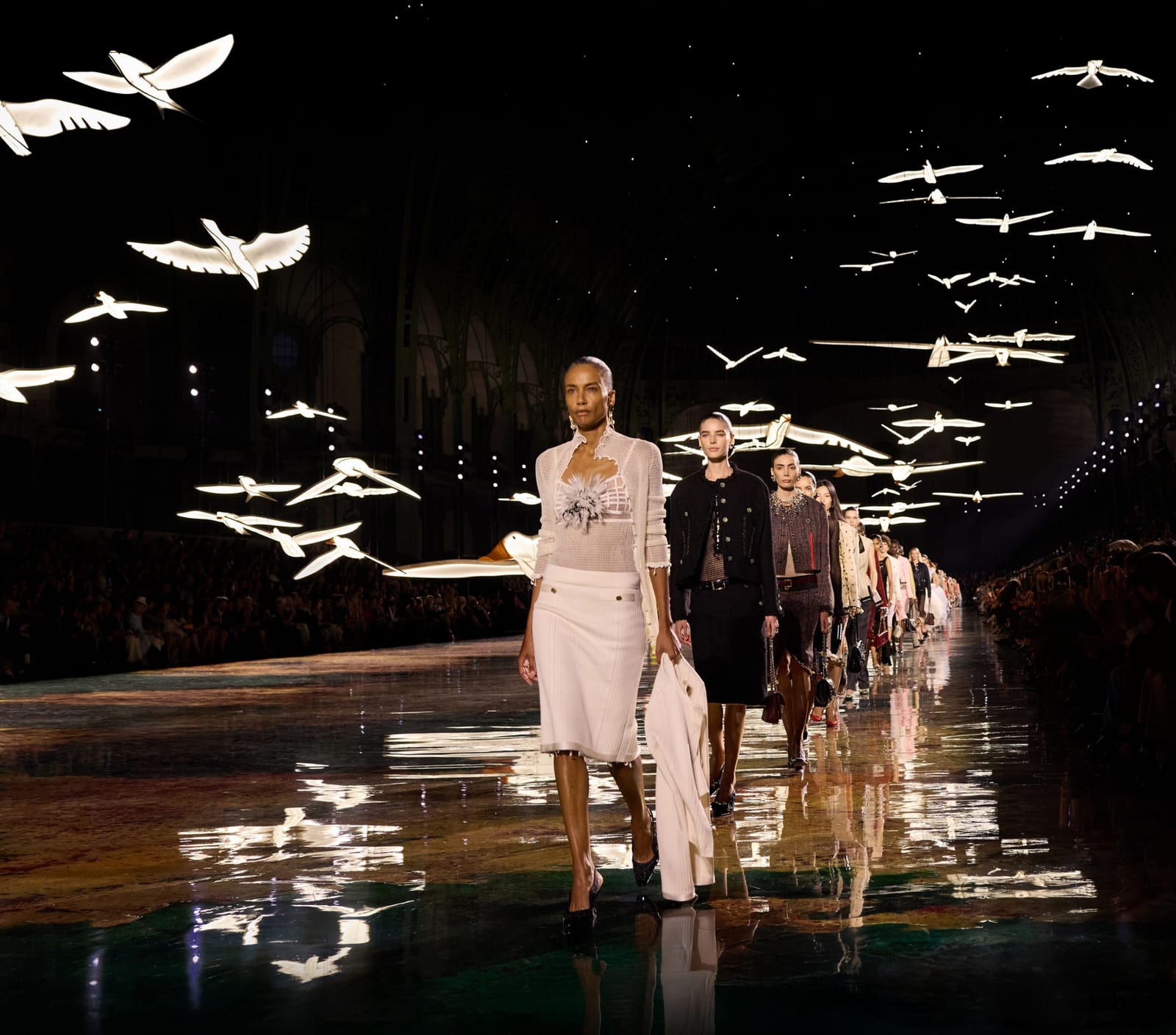 The Chanel Spring 2027 finale at the Grand Palais: a model in a sheer white knit cardigan over a bralette with a grey flower, and a white pencil skirt, carrying a white jacket, leads a line of models in tweed jackets and skirts under glowing white birds, on a reflective floor