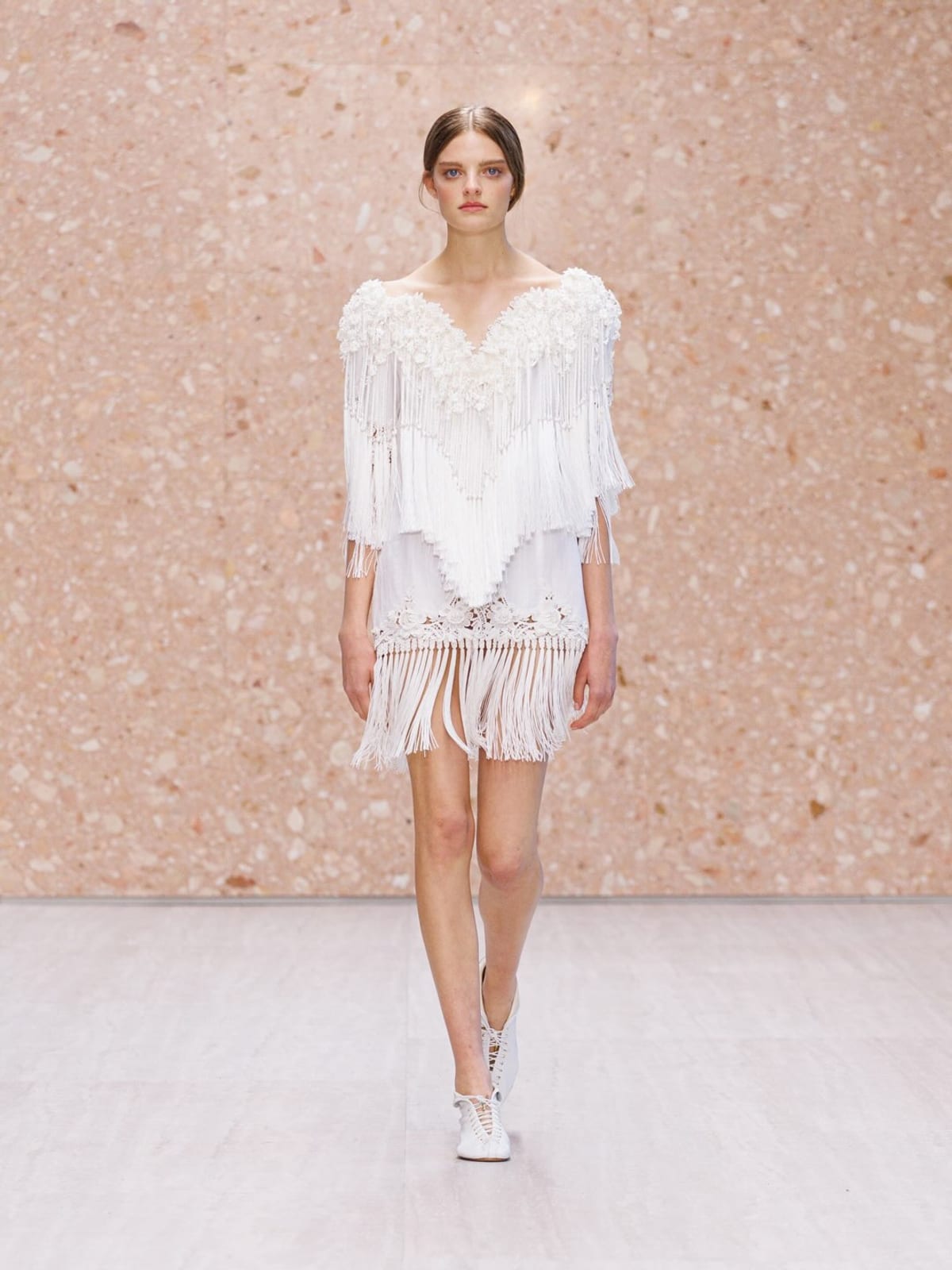 A Chloé Spring/Summer 2027 look on a pink terrazzo-walled runway: a white off-the-shoulder fringed top with a plunging neckline over a fringed white mini skirt