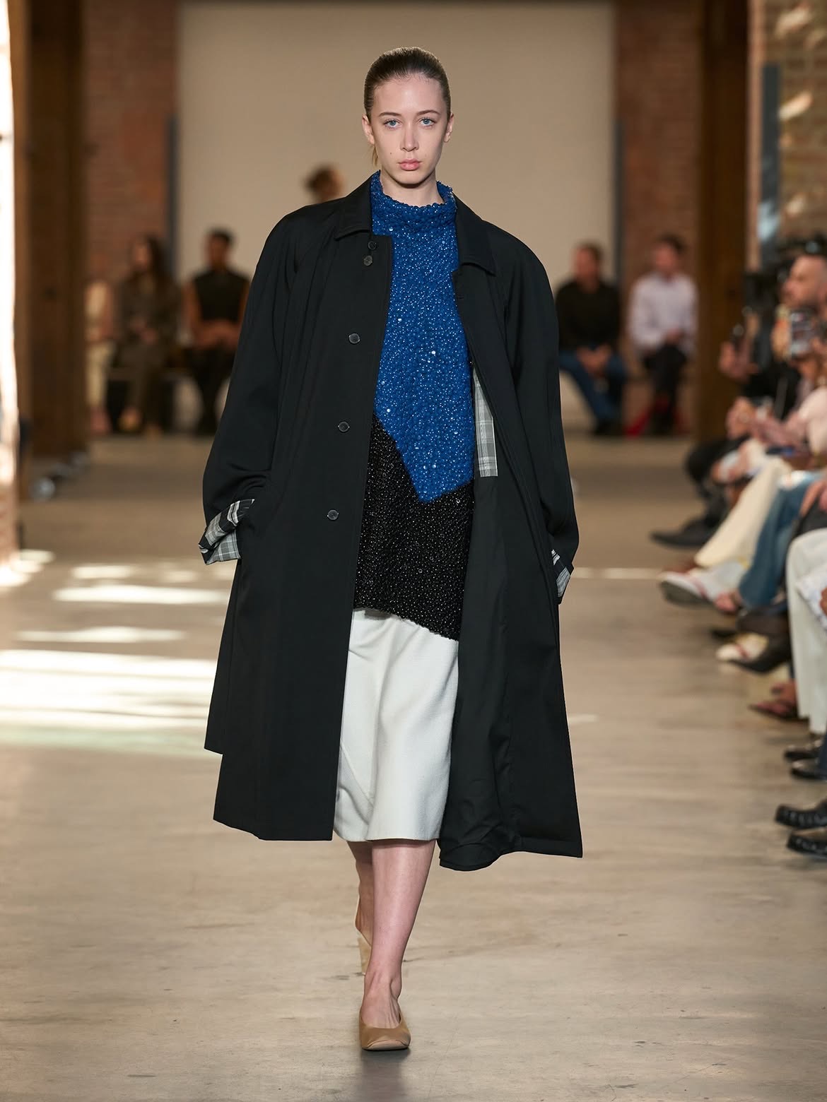 A model in a long black coat with a plaid lining, over a sequined blue top and an ivory skirt