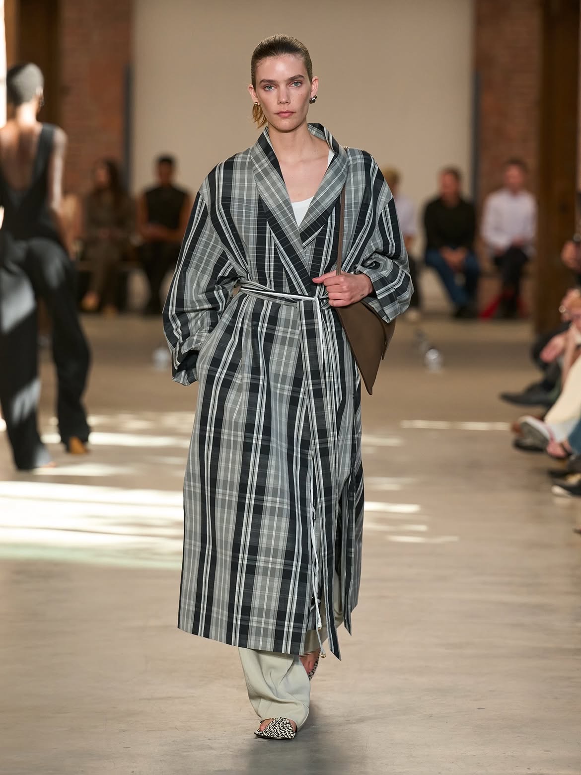 A model in a long grey and black plaid coat tied at the waist, over ivory trousers