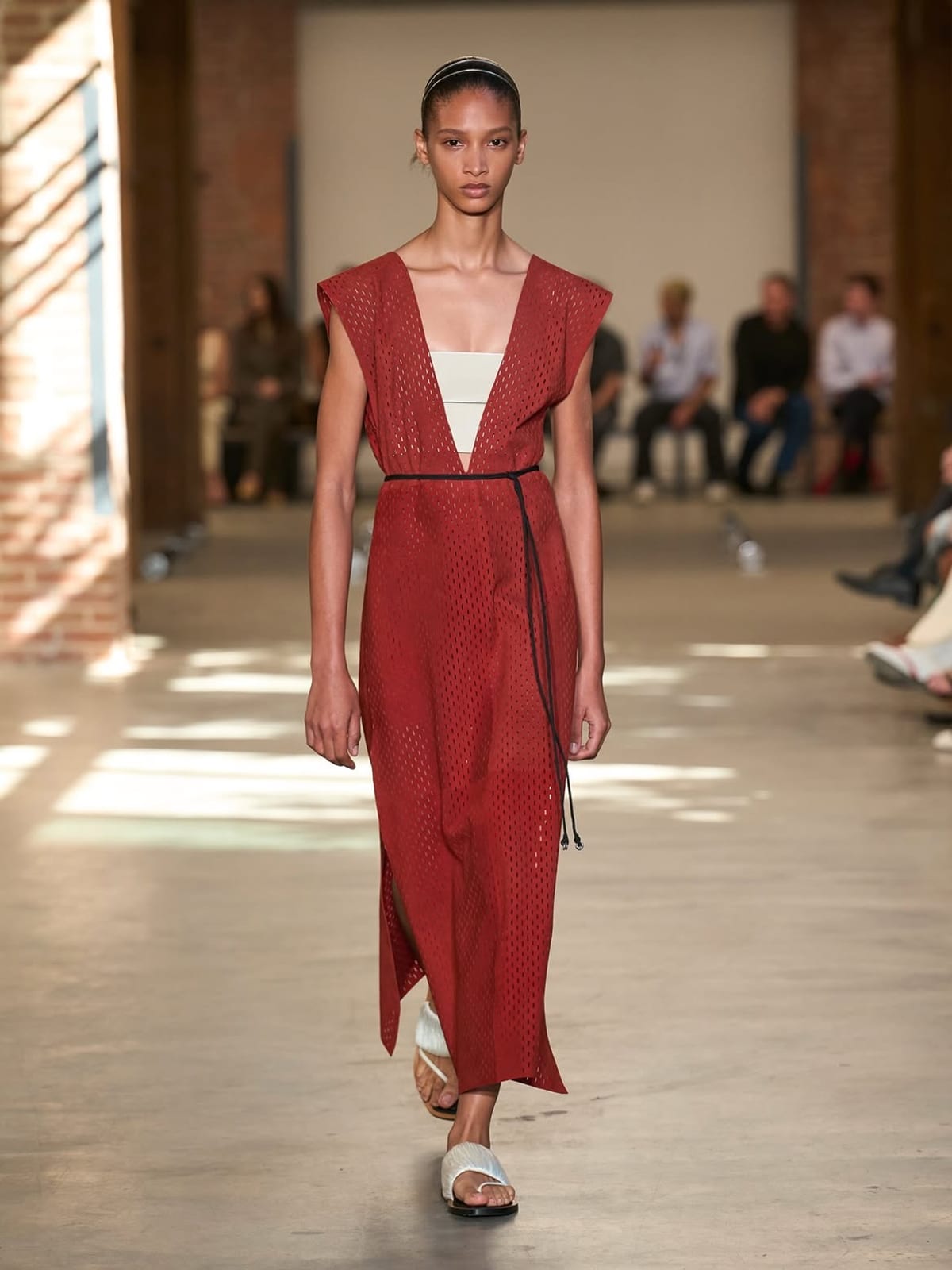 A model in a rust-red perforated sleeveless dress with a deep V-neck over a white bandeau, tied with a thin black cord belt