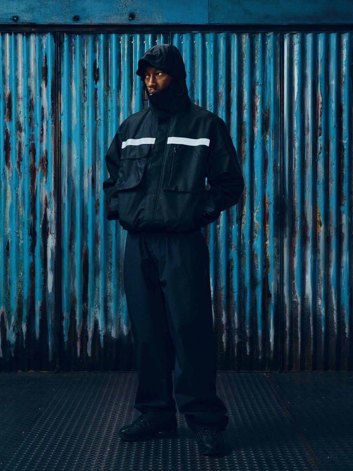 A model in Corteiz's black 'It Ain't Safe' Litework jacket and matching trousers, hood up, from the Konnichiwa 10th anniversary collection