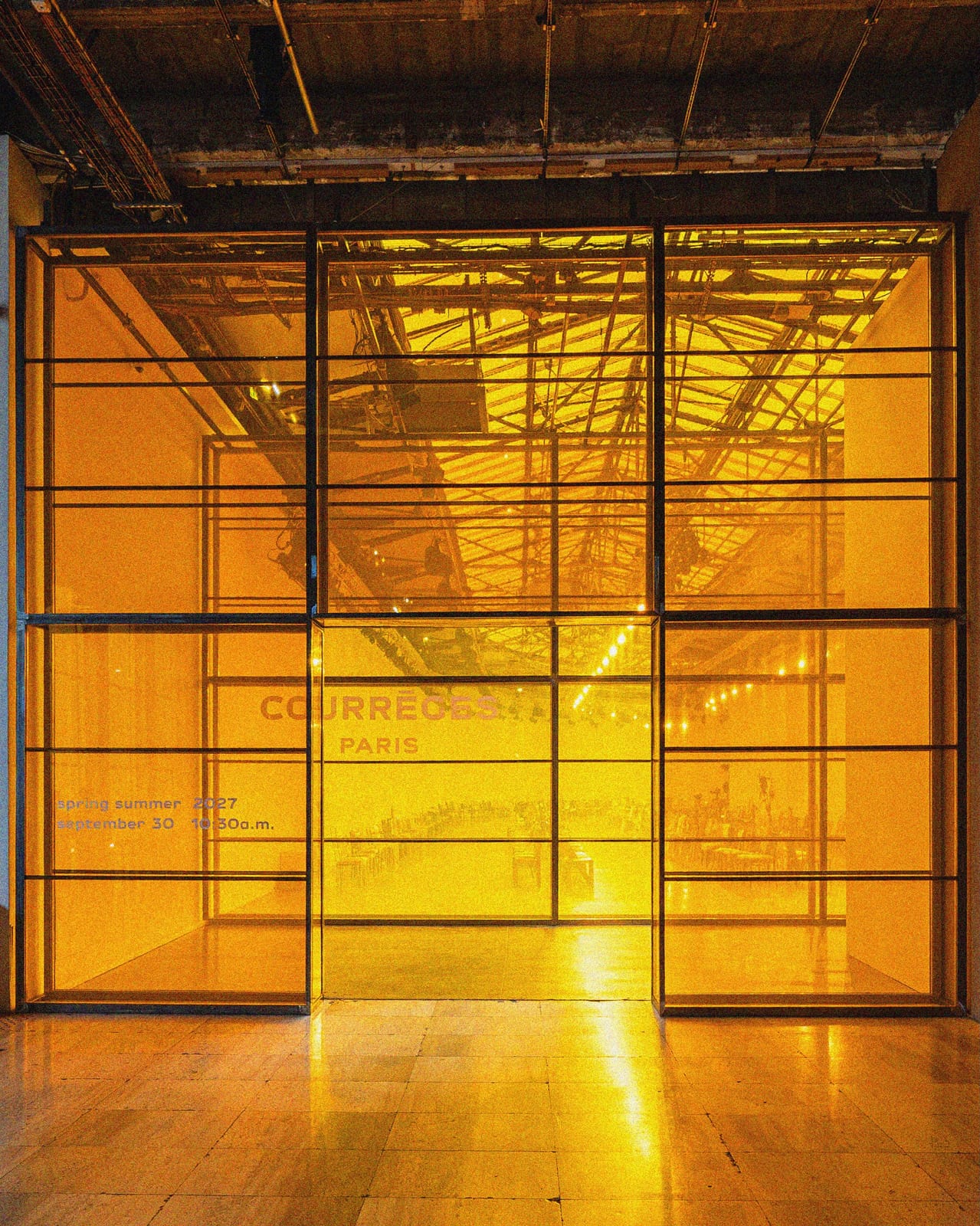 The entrance to the Courrèges Spring/Summer 2027 show: a glass-and-steel frame lit amber, with the Courrèges Paris name and the show date and time printed on the glass