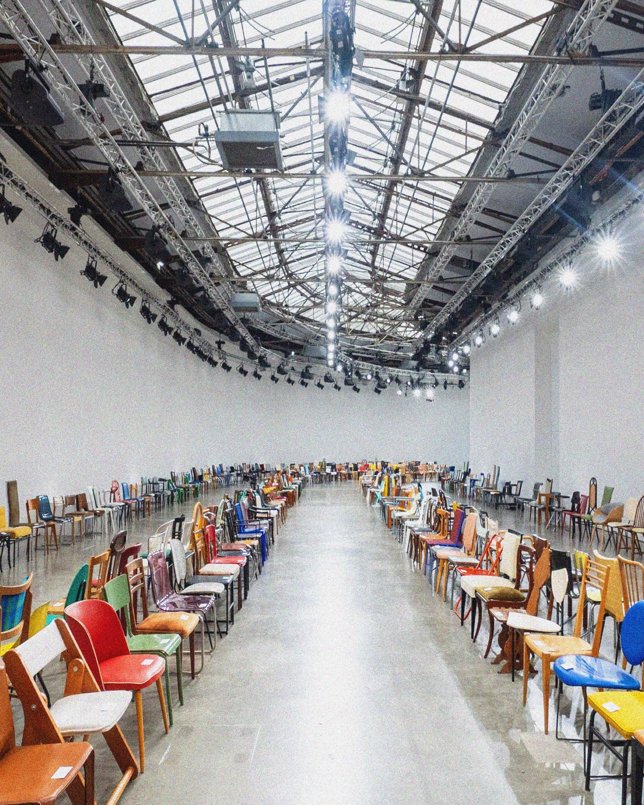 The Courrèges Spring/Summer 2027 show space at the Palais de Tokyo: two long rows of mismatched, multicoloured chairs under a glass roof, around a bare concrete runway