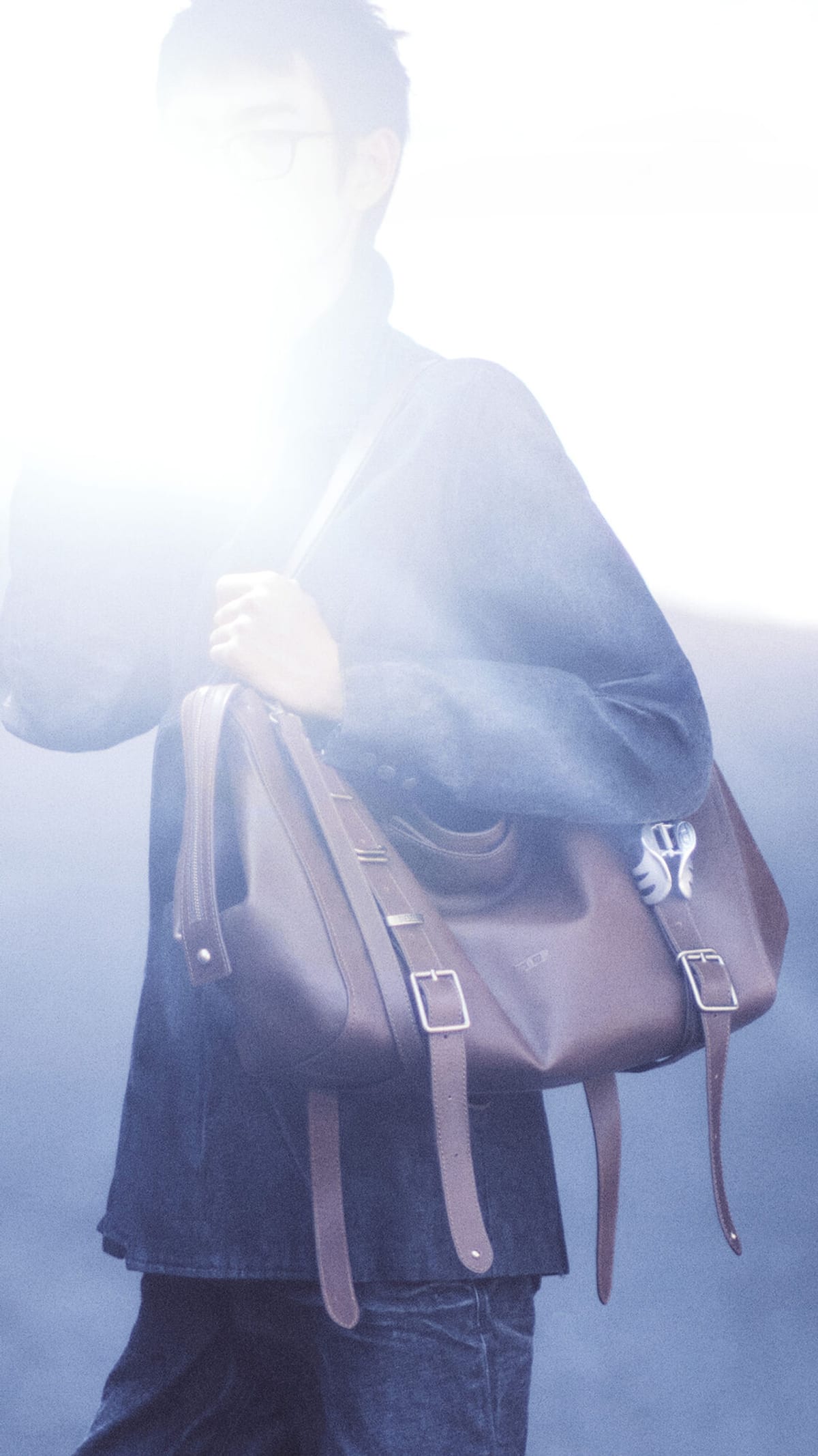 A Diesel campaign image: a person in a dark denim jacket, their face lost in a flare of pale light, carrying the brown leather D-One bag on one shoulder, its buckled straps hanging down