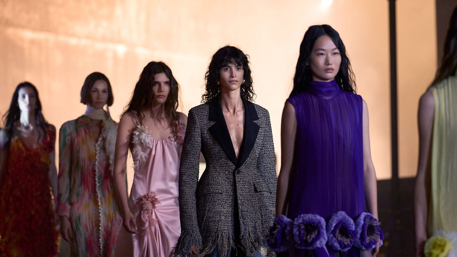 Models in Dior's Cruise 2027 collection lined up at the May show at LACMA: a pink satin slip dress, a black-lapelled tweed jacket with a fringed hem, and a high-necked purple pleated dress with large purple flowers at the hem