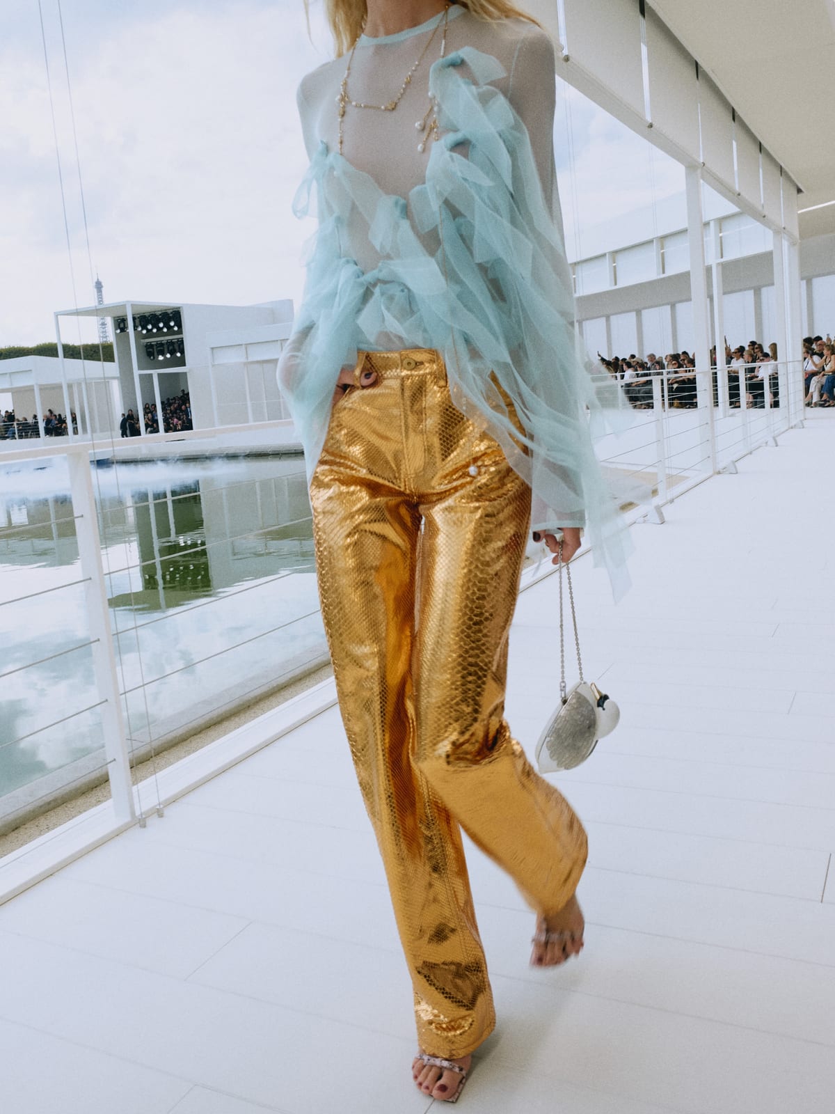 A Dior Summer 2027 look on the white catwalk over the Tuileries basin: metallic gold trousers under a sheer, ruffled pale-blue top