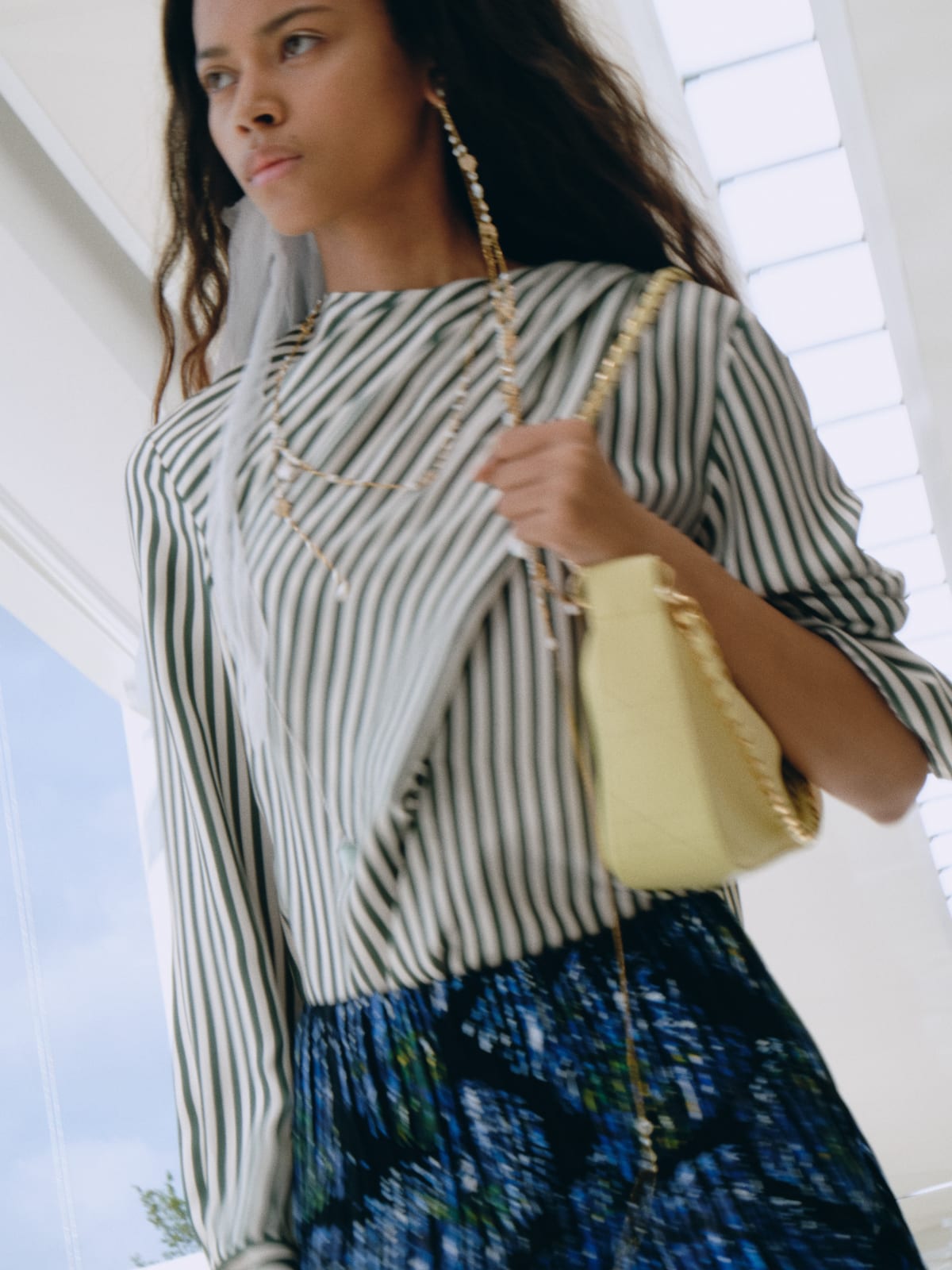 A Dior Summer 2027 look: a green-and-white striped shirt over a dark patterned skirt, with a small yellow chain bag