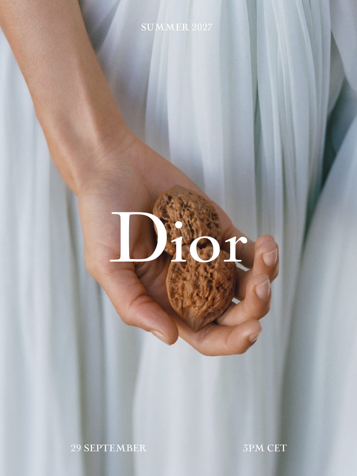 Dior's Summer 2027 invitation: a hand holding a walnut against pale pleated fabric, with the show date, 29 September
