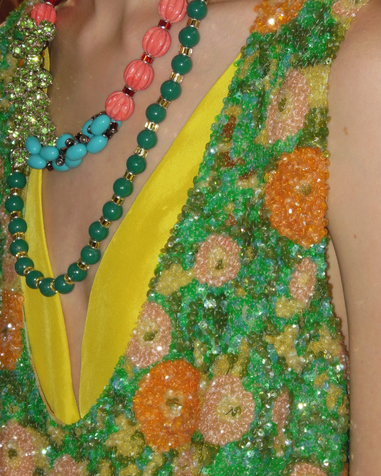 Close-up of a Dries Van Noten Spring/Summer 2027 look: a deep V-neck vest densely embroidered in green, orange and peach sequin flowers over a bright yellow top, with long coral, teal and turquoise bead necklaces