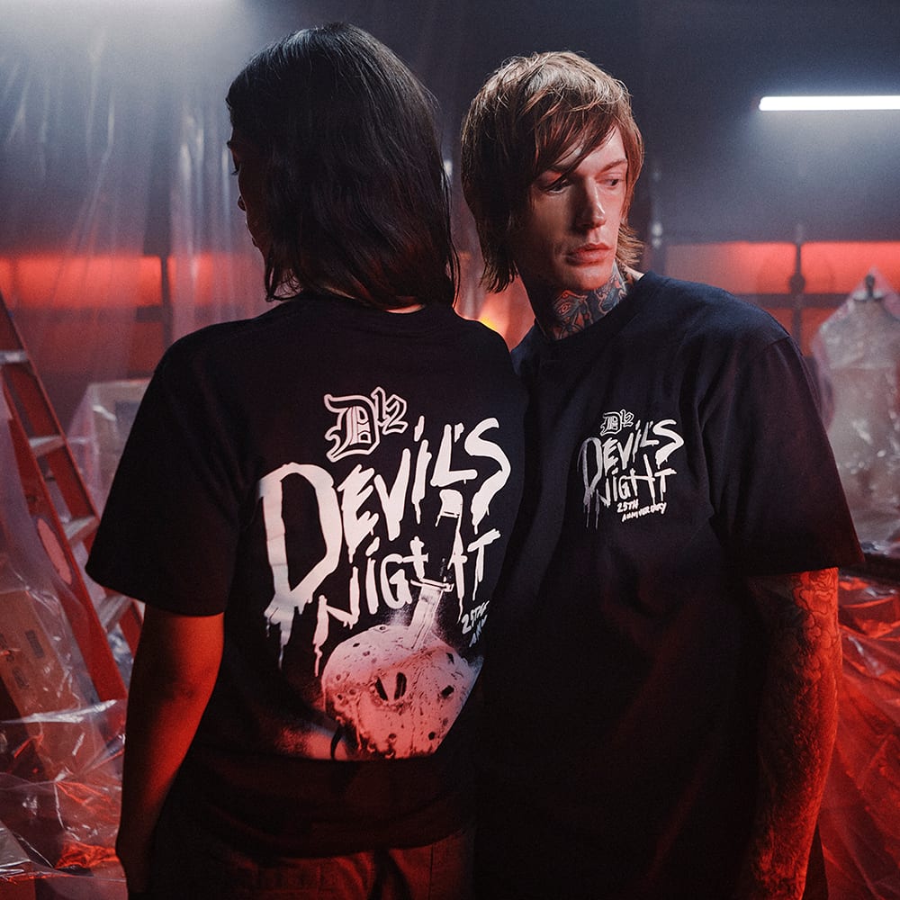 Two people in black Devil's Night 25th anniversary tees among plastic sheeting in a red-lit warehouse, one seen from behind with the large Devil's Night and hockey mask print across the back, the other wearing the small chest print