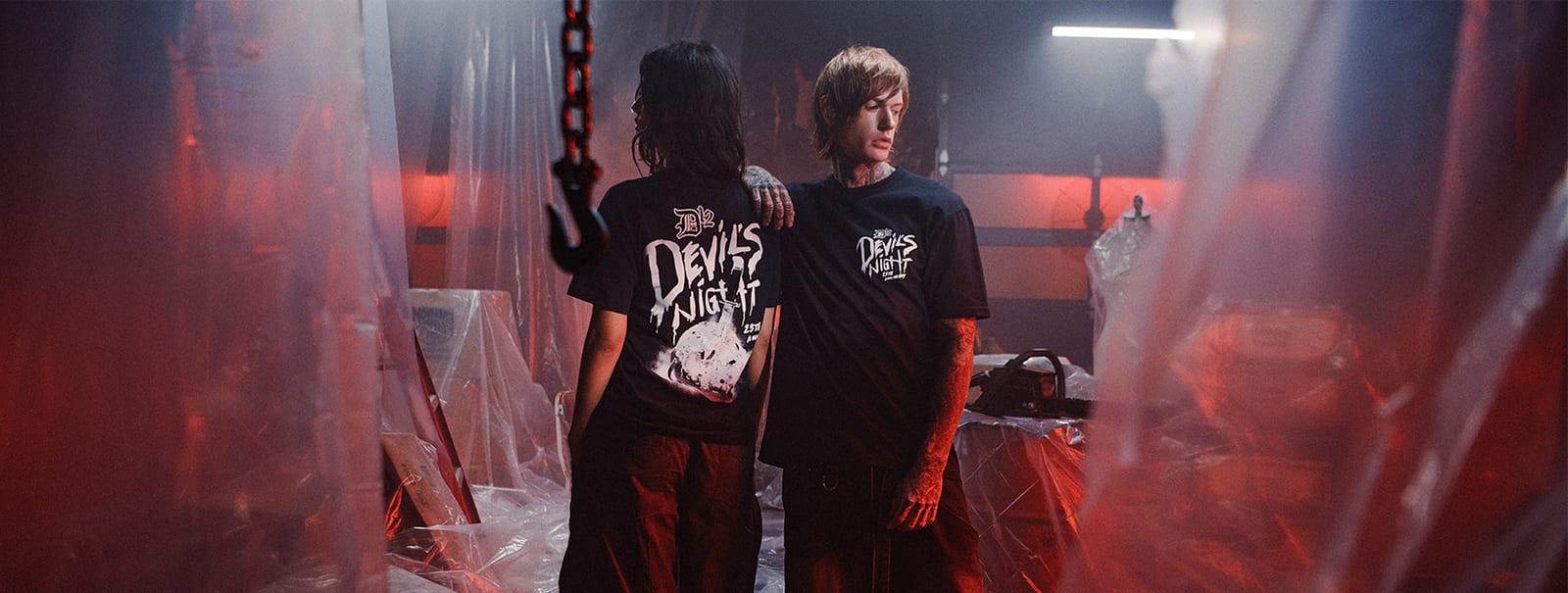 Two people in black Devil's Night 25th anniversary tees standing among hanging plastic sheeting in a red-lit warehouse, one seen from behind with a large Devil's Night print across the back