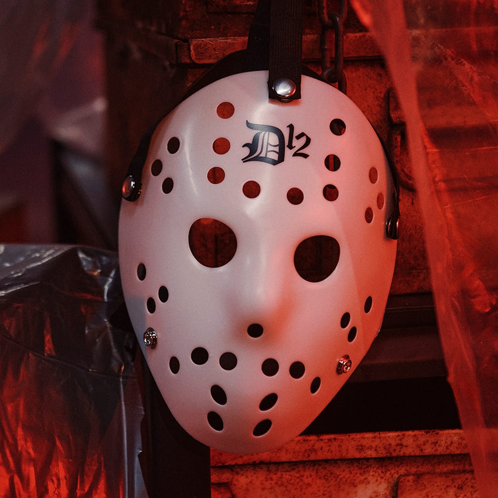 A white hockey goalie mask with rows of breathing holes and a black D12 logo above the eye holes, hanging on a strap against red-lit metal crates