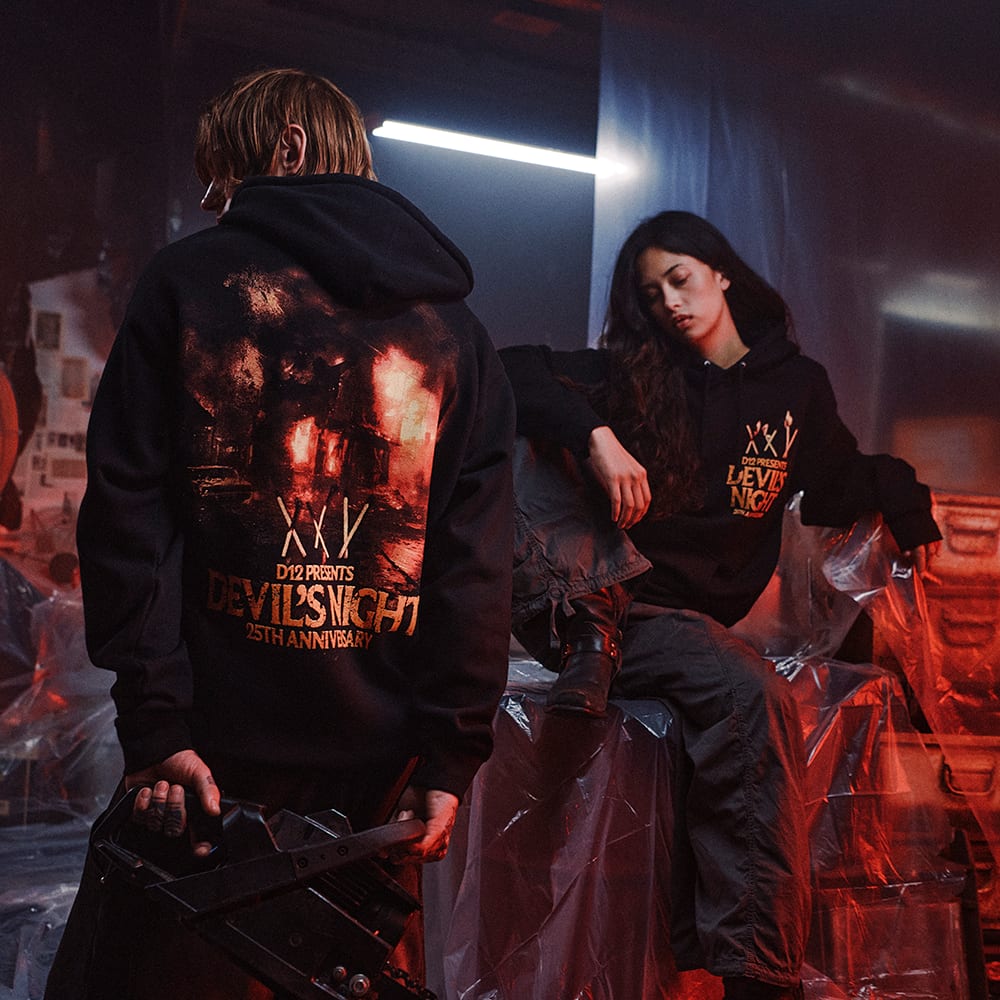 A man seen from behind in a black hoodie with a large printed Devil's Night artwork across the back, beside a seated woman in the same hoodie showing its small chest print of crossed match sticks and the words D12 Presents Devil's Night 25th Anniversary, in a red-lit room