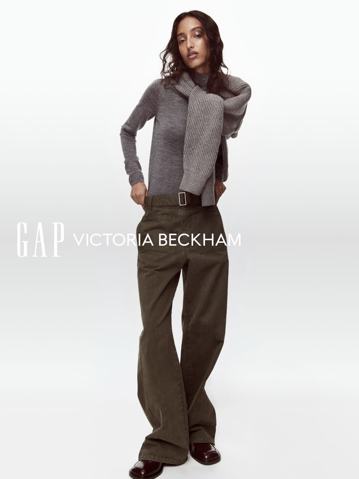 Mona Tougaard in a fine grey knit with a ribbed grey sweater tied over her shoulders, and wide olive-brown trousers with a belt, against a white backdrop with the Gap and Victoria Beckham logos