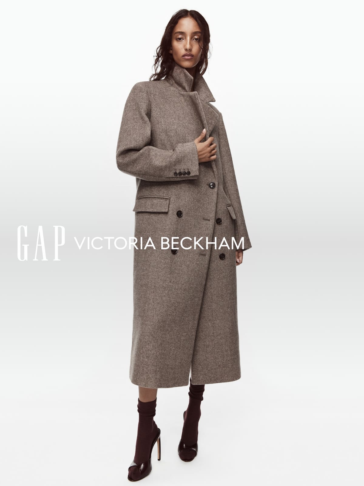 Mona Tougaard in a long double-breasted coat in flecked taupe wool, worn with burgundy socks and open-toe heels, against a white backdrop with the Gap and Victoria Beckham logos