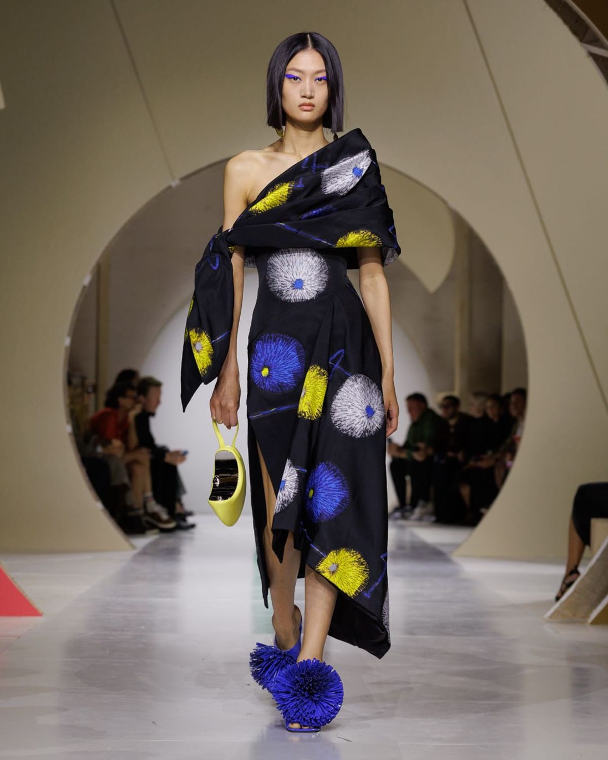 A model in an asymmetric black dress with an oversized knotted off-the-shoulder sash, printed with large blue, yellow and white starburst flowers, with blue pom-pom sandals and a yellow handbag