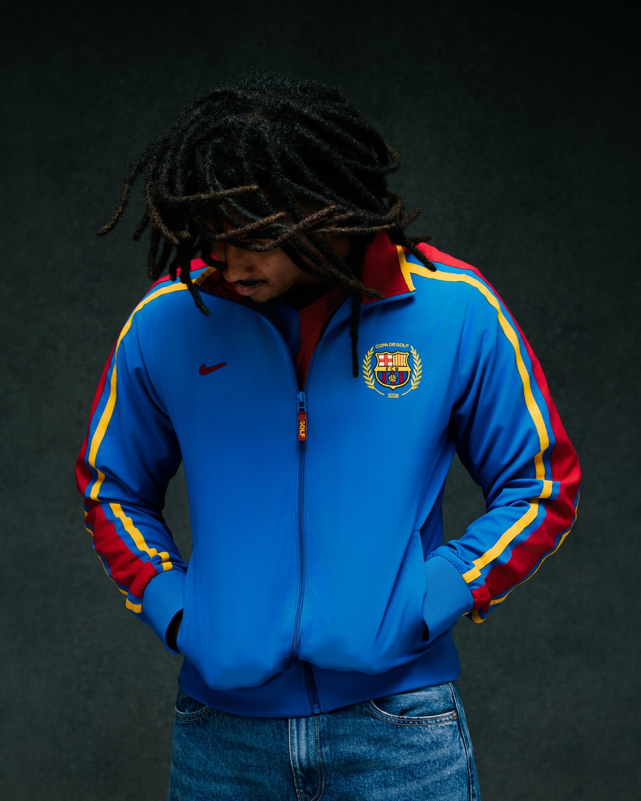 Jules Koundé, head bowed with his locs falling over his face, in GOLF WANG's royal blue Copa De Golf track jacket and blue jeans, with red, yellow and blue striped sleeves, a dark red Nike Swoosh, a GOLF zipper pull and an FC Barcelona crest inside a laurel wreath reading Copa De Golf 2026