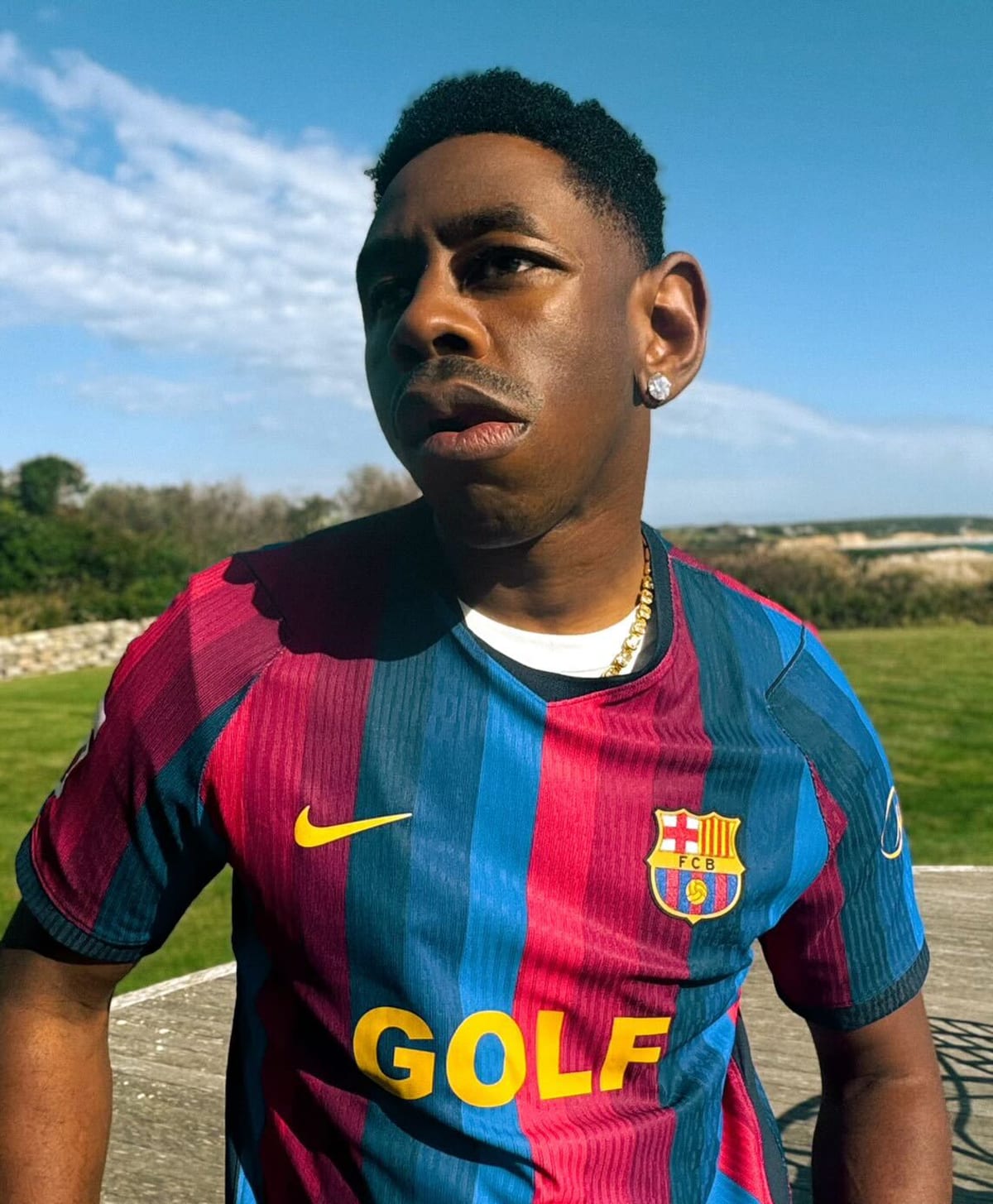 Tyler, The Creator outdoors under a blue sky in FC Barcelona's red and blue striped shirt, with a yellow Nike Swoosh, the club crest and a yellow GOLF logo across the chest