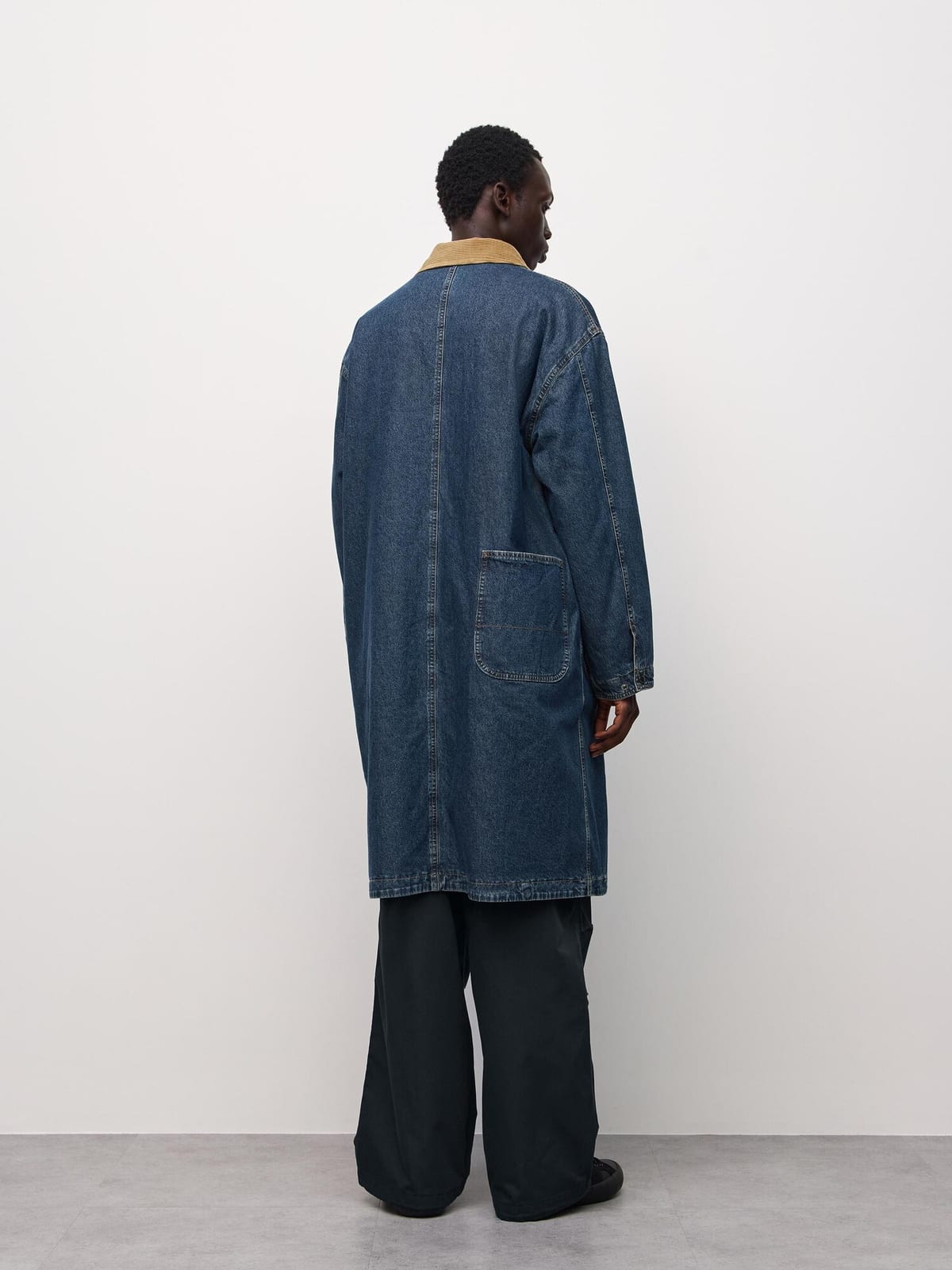 The back of the long blue denim work coat, with a tan corduroy collar and a patch pocket at the hip, worn by a man in wide black trousers against a white wall